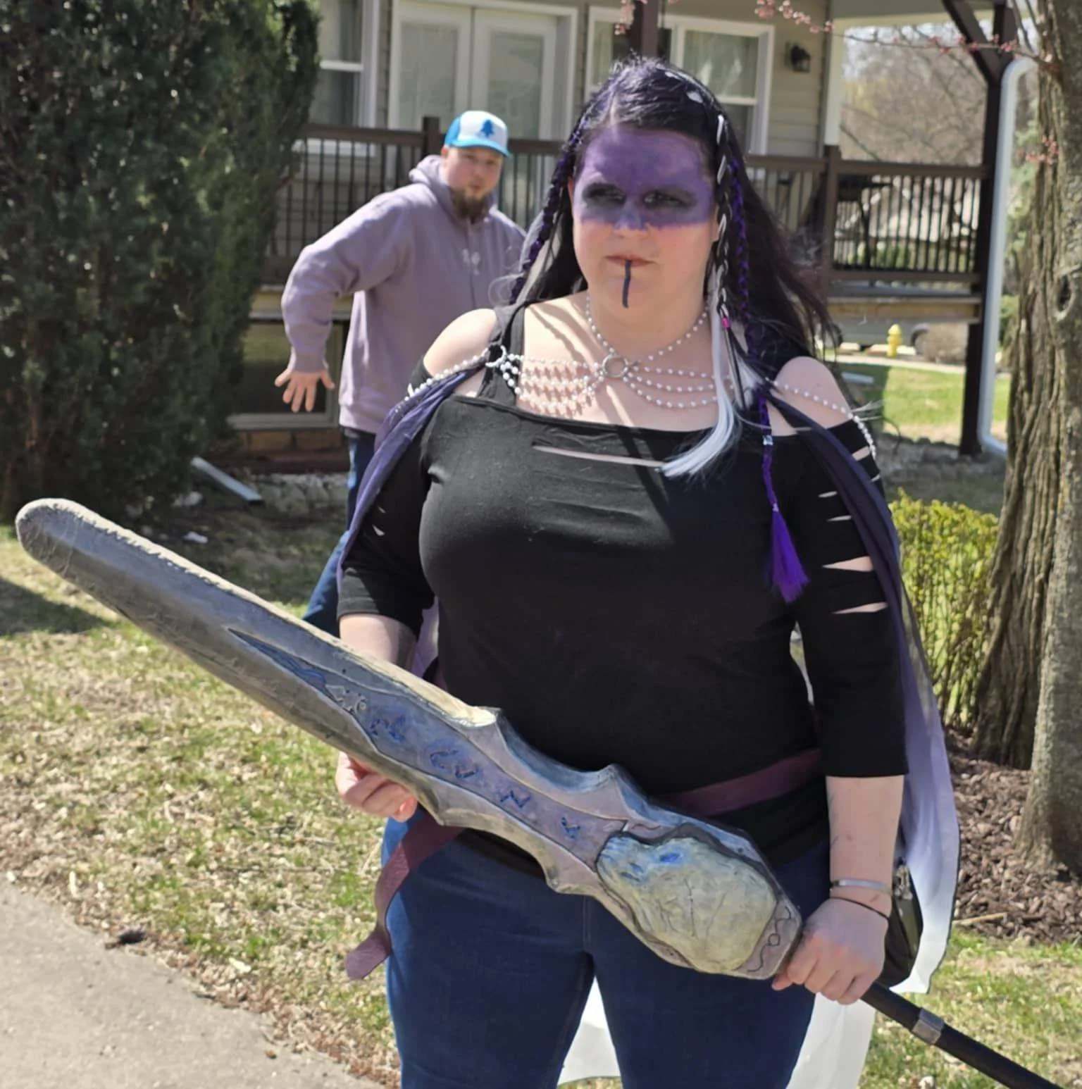 A woman dressed as a fantasy warrior with purple face paint, dark purple hair with white and purple braids, wearing a black cut-out top, pearl necklaces, and holding a large fake sword, standing outdoors with a man in a hoodie and cap in the background near a house and trees.