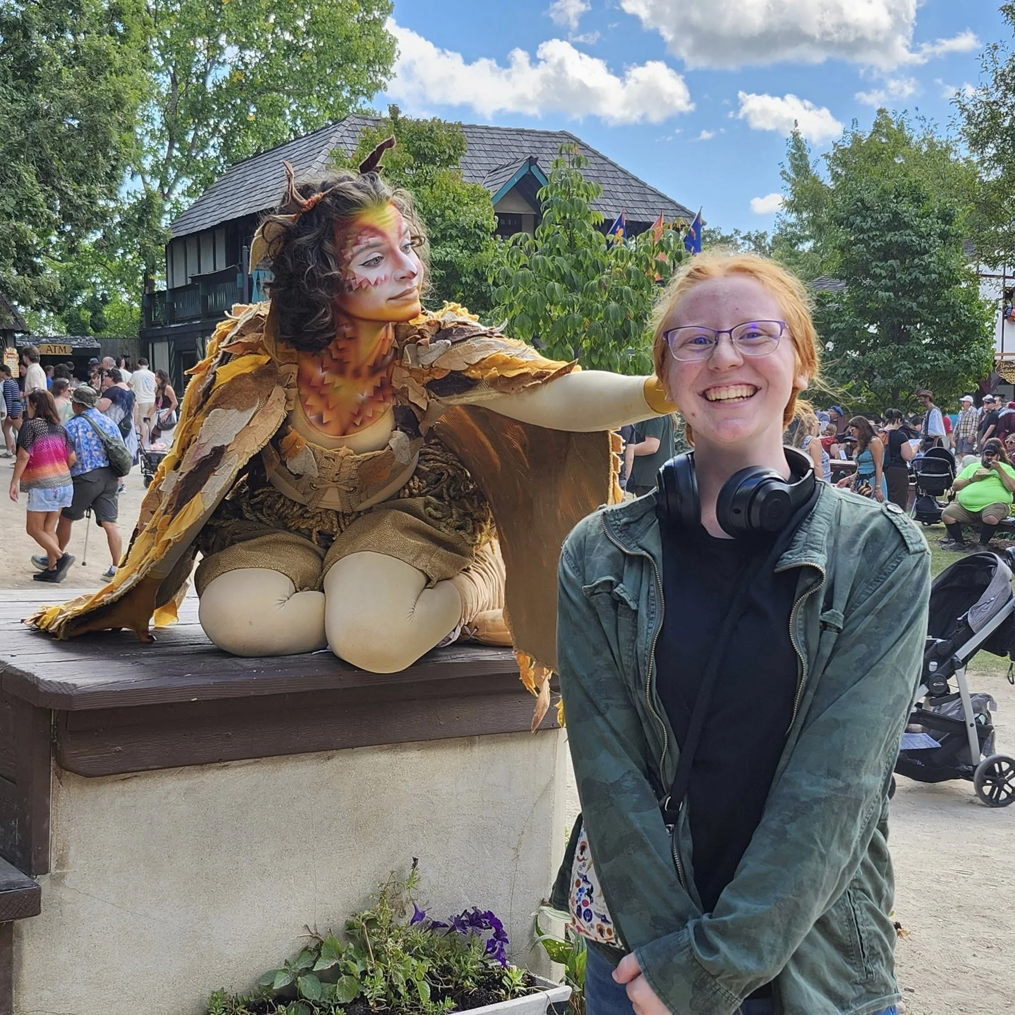 A smiling person with short red hair, glasses, and headphones around their neck stands in front of a fantasy character dressed in an elaborate costume at an outdoor event.