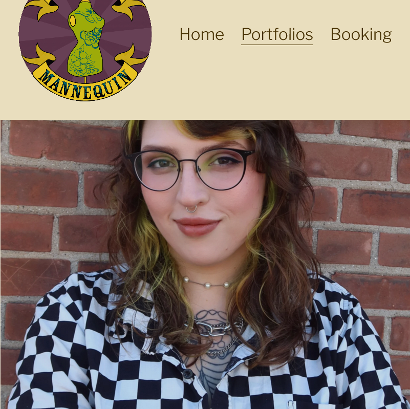 A young woman with wavy brown hair and green highlights, wearing glasses, a septum piercing, and layered necklaces, standing in front of a brick wall.