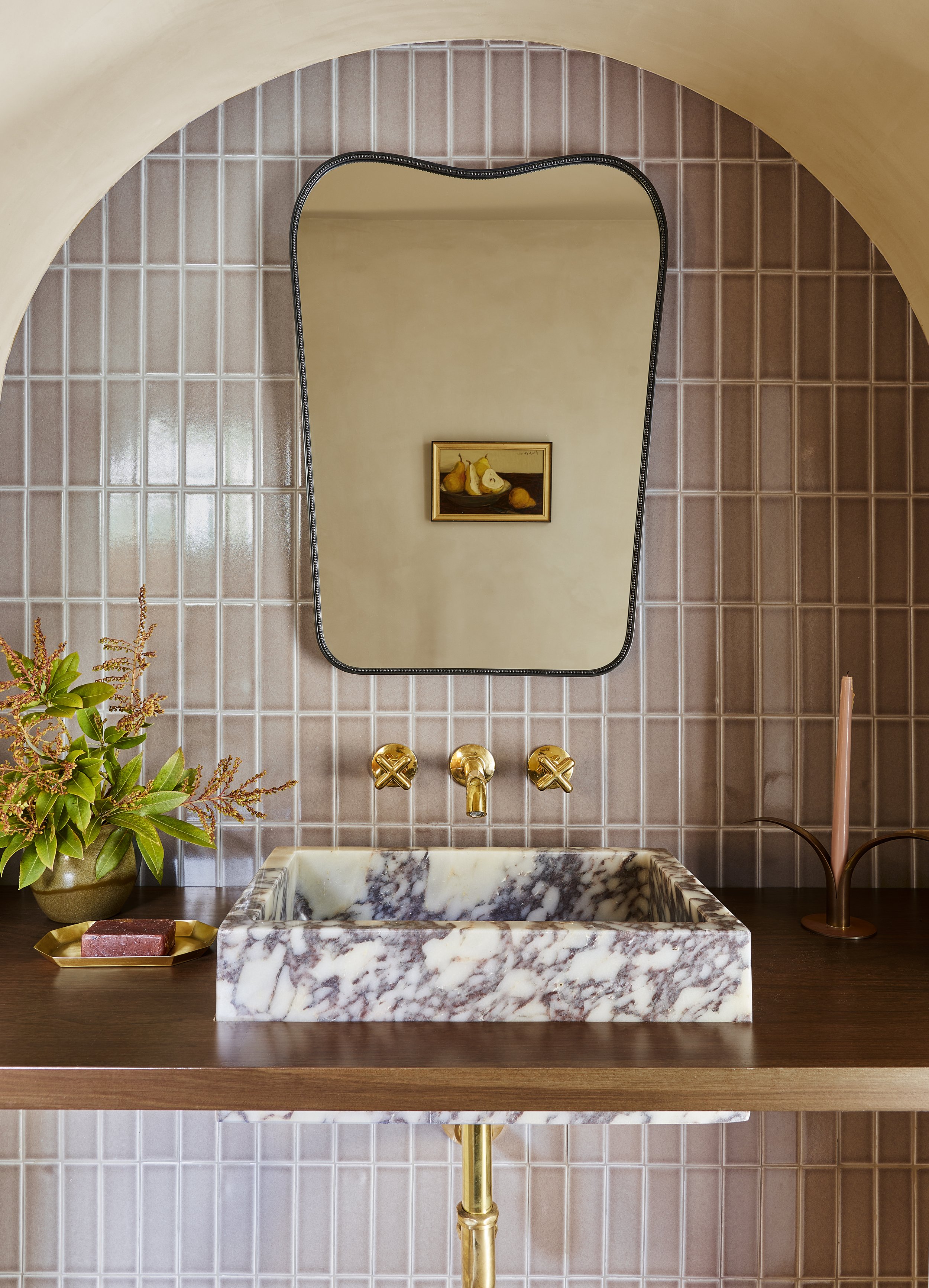 A bathroom vanity area featuring a marble sink, gold faucet, a mirror with a black frame, pink tiled wall, green plant, and a small gold tray with soap.