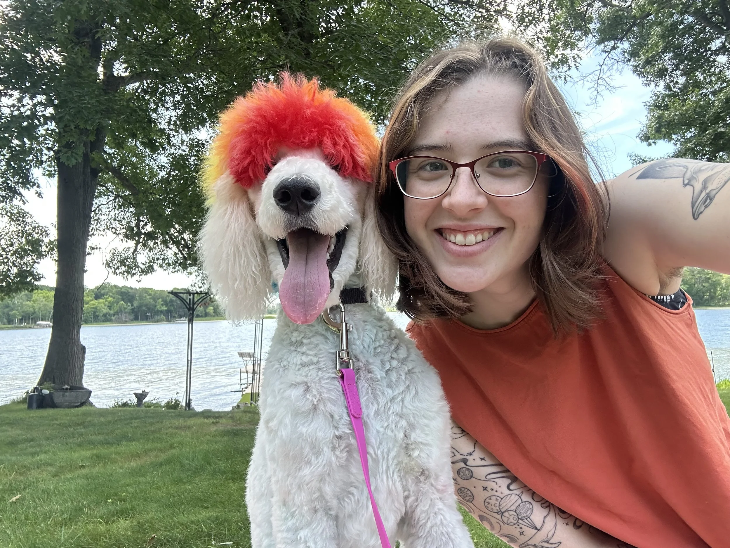 A smiling woman with glasses and a necklace is taking a selfie with a dog that has colorful markings on its fur. The dog is wearing a pink hood and has blue and purple paint or markings on its face, ears, and body. The dog is standing on a grooming table covered with a blue towel in a pet grooming or veterinary clinic.