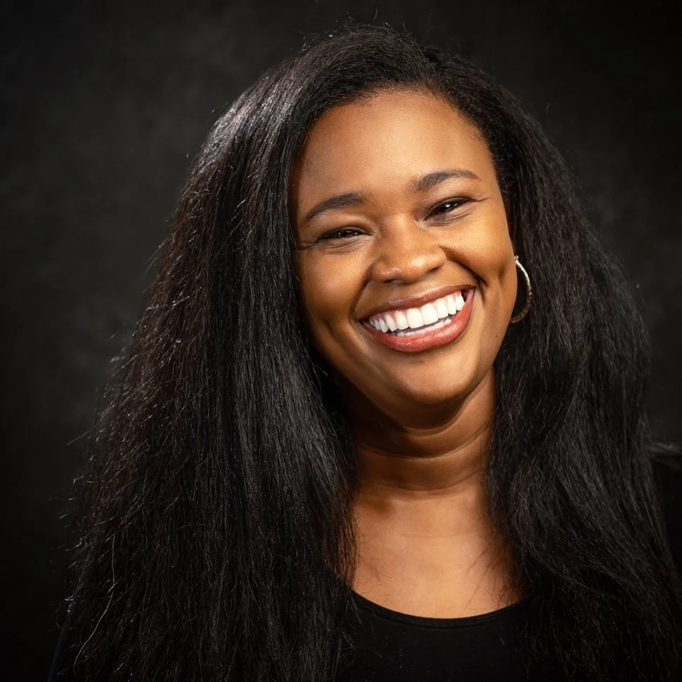 Jess Moore Matthews, a smiling Black woman with long black hair. She is wearing a black shirt and gold hoop earrings.