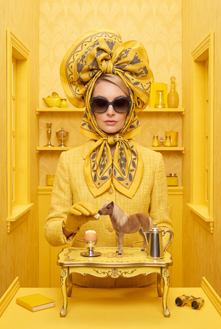 Model wearing sunglasses and an oversized yellow patterned silk scarf headpiece beside a miniature horse, egg and silver teapot.