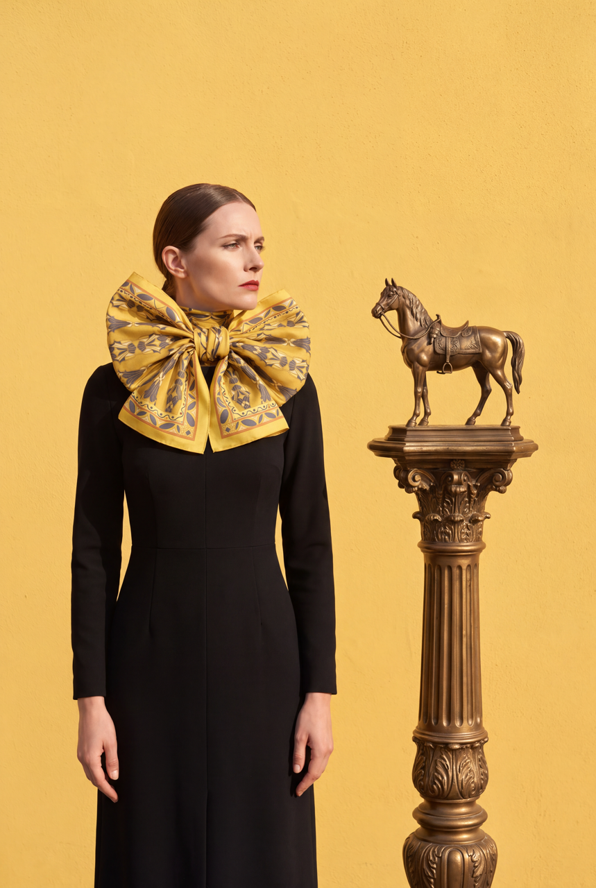 Model in a black dress wearing an oversized yellow patterned silk scarf bow beside a bronze horse statue on a decorative pedestal.
