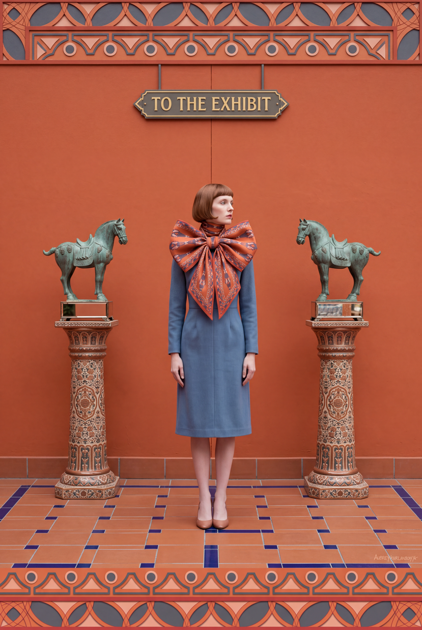 Model wearing a blue-grey coat and oversized orange patterned silk scarf bow between two decorative horse statues against an orange wall.