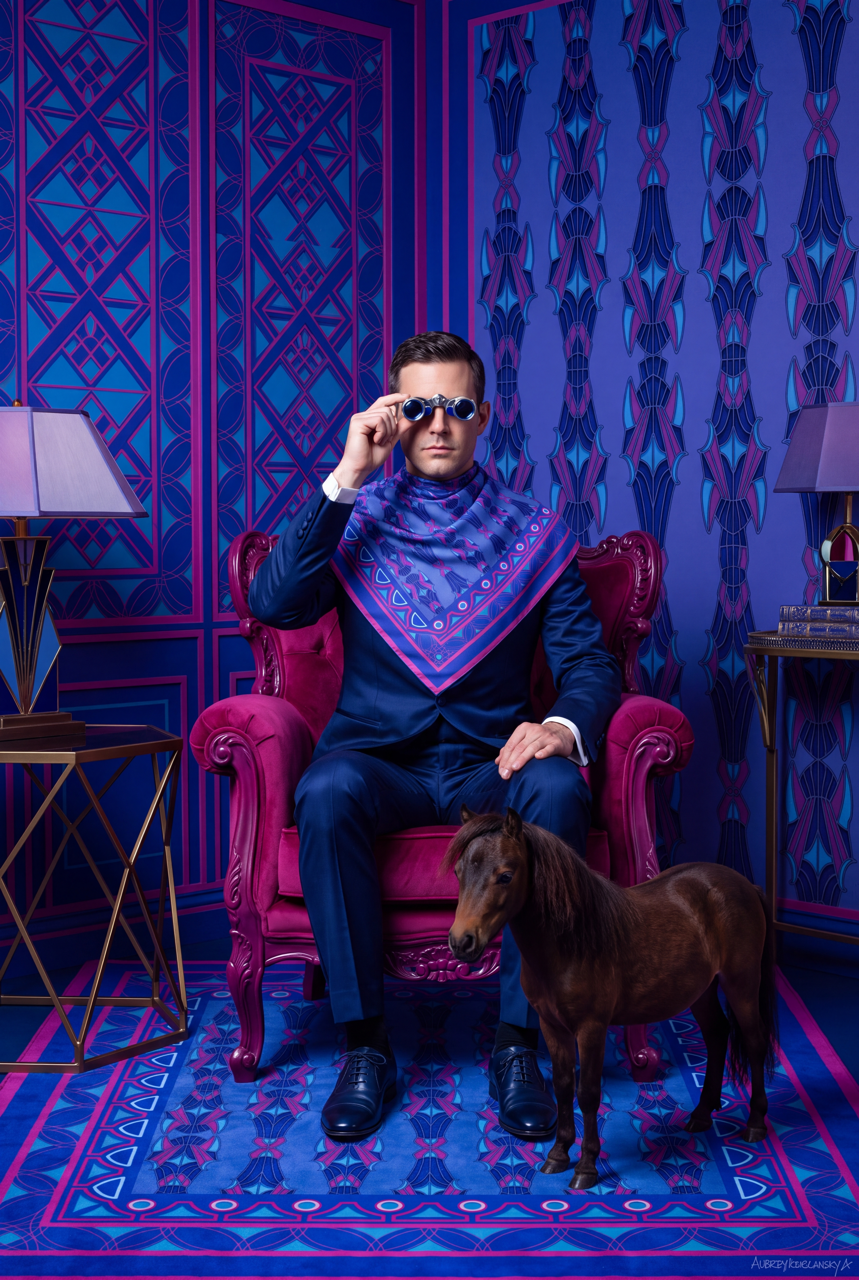 Model in a blue suit wearing a blue-and-magenta patterned silk scarf, seated in a magenta armchair beside a miniature horse.