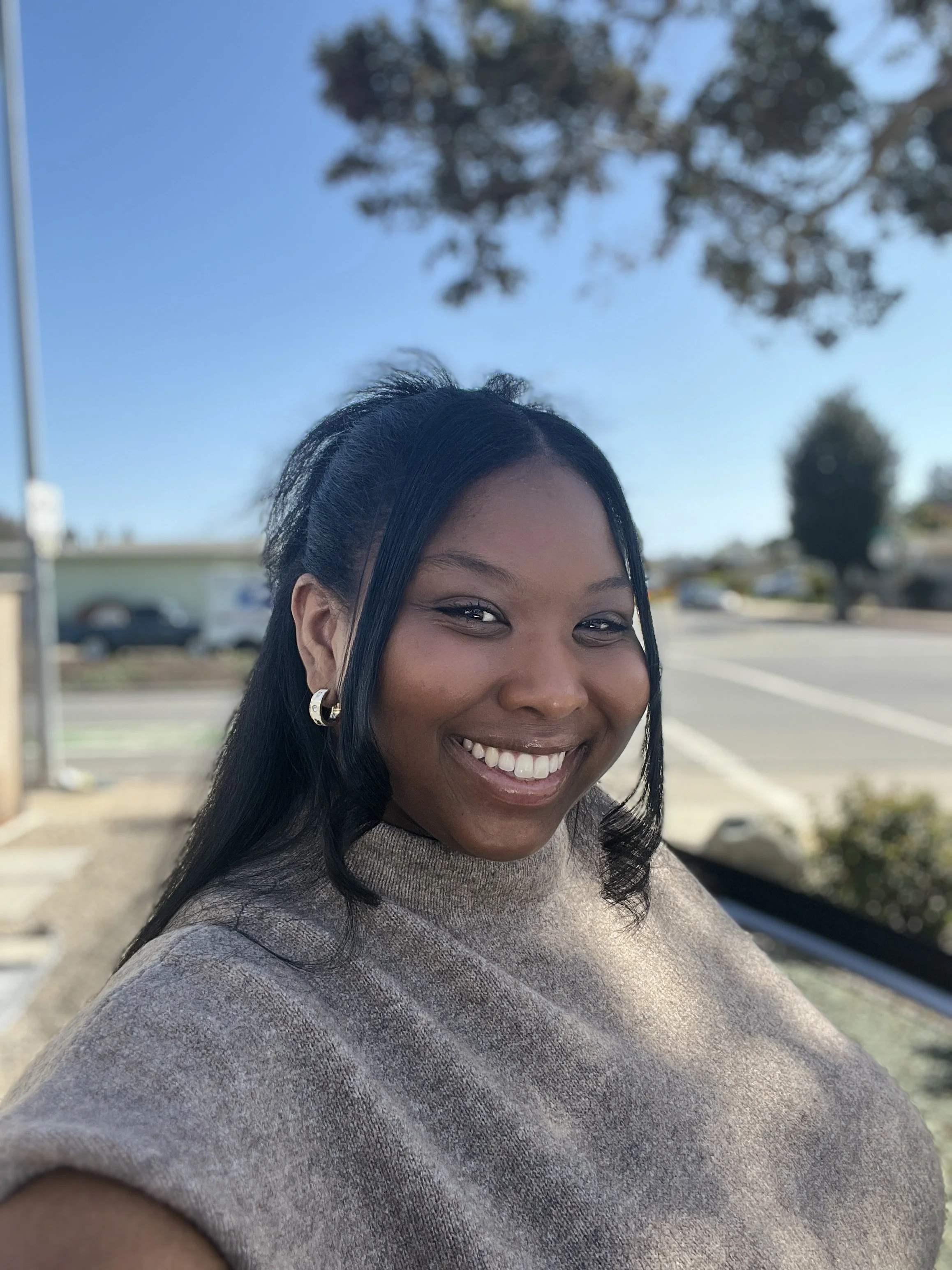 A young woman with dark hair smiling outdoors on a sunny day, wearing a beige top and earrings.