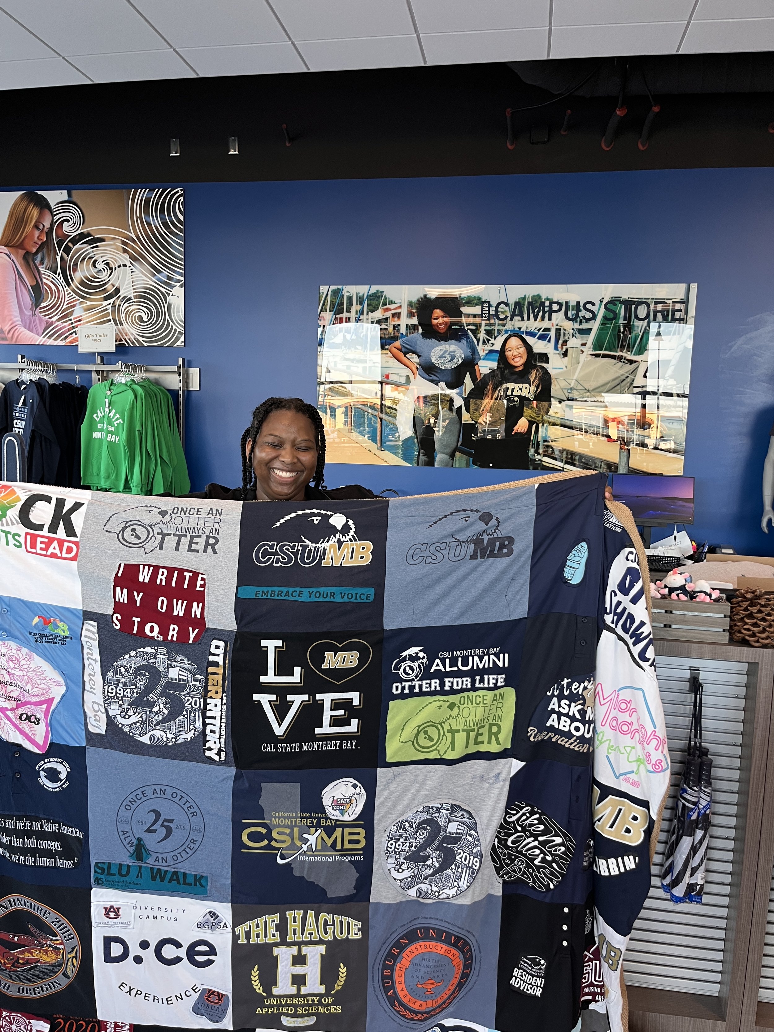 Woman smiling and holding a quilt with various university and college logos and slogans, standing inside a store with merchandise in the background.