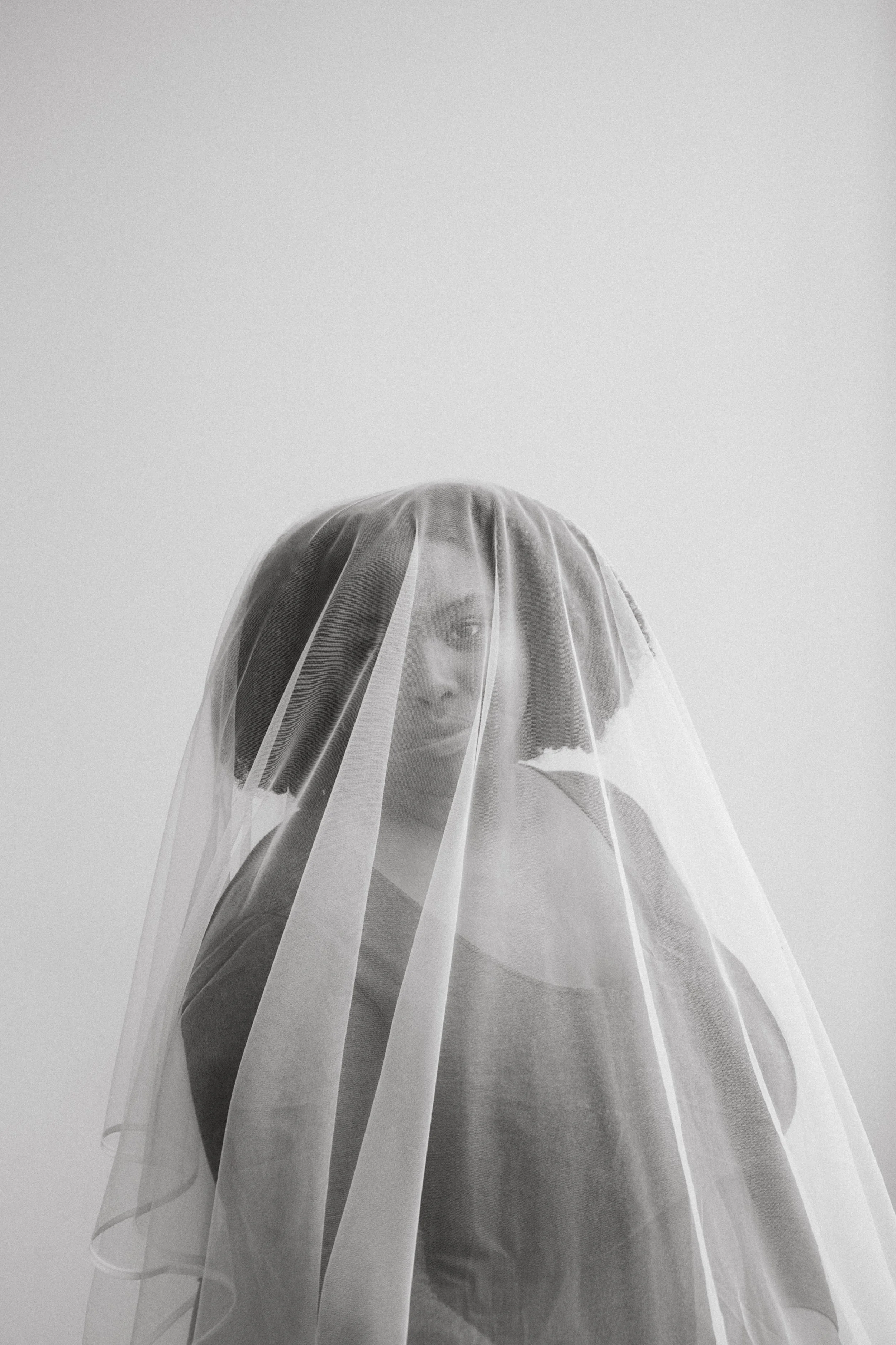 A woman with short hair behind a sheer veil, partially obscuring her face, in black and white.