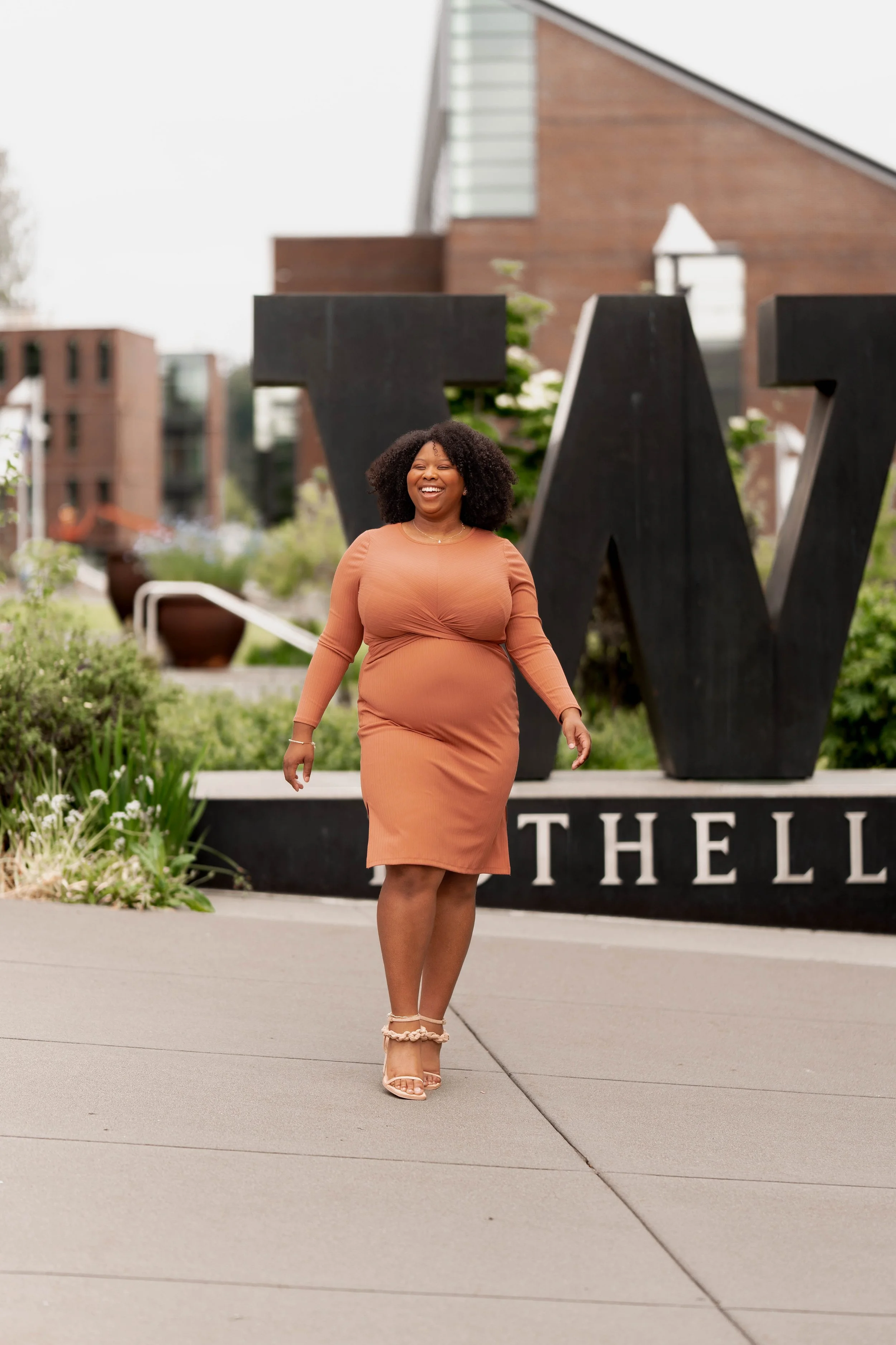 A woman in a tan dress walking and smiling in front of large black letters that spell 'W' and 'THELL' at an outdoor location with greenery and modern buildings in the background.