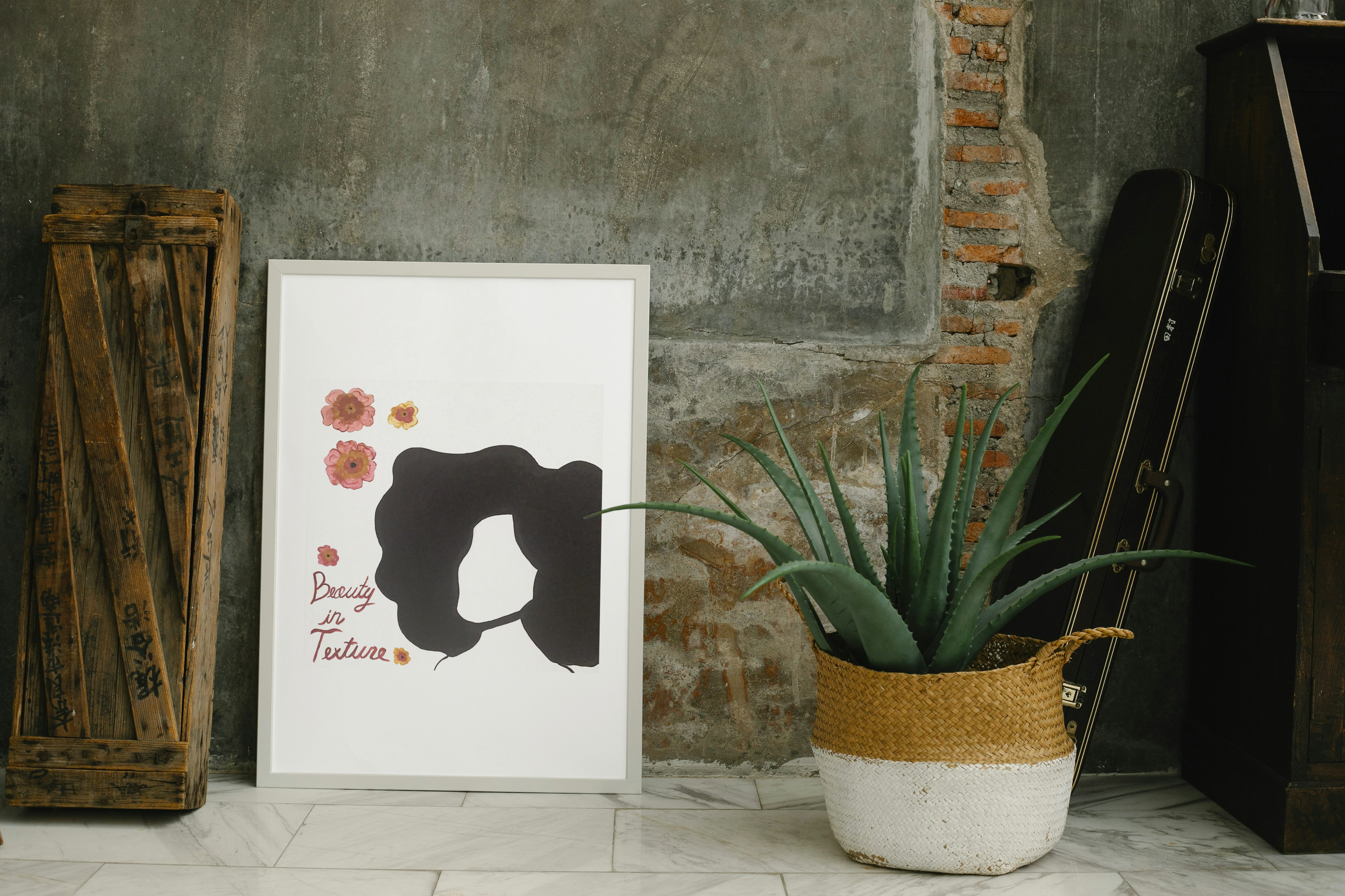 Interior decor with a framed poster featuring a black silhouette of a woman's face with flower embellishments and the text 'Beauty in Texture', a potted aloe vera plant in a woven basket, and a vintage wooden crate leaning against a distressed concrete wall.