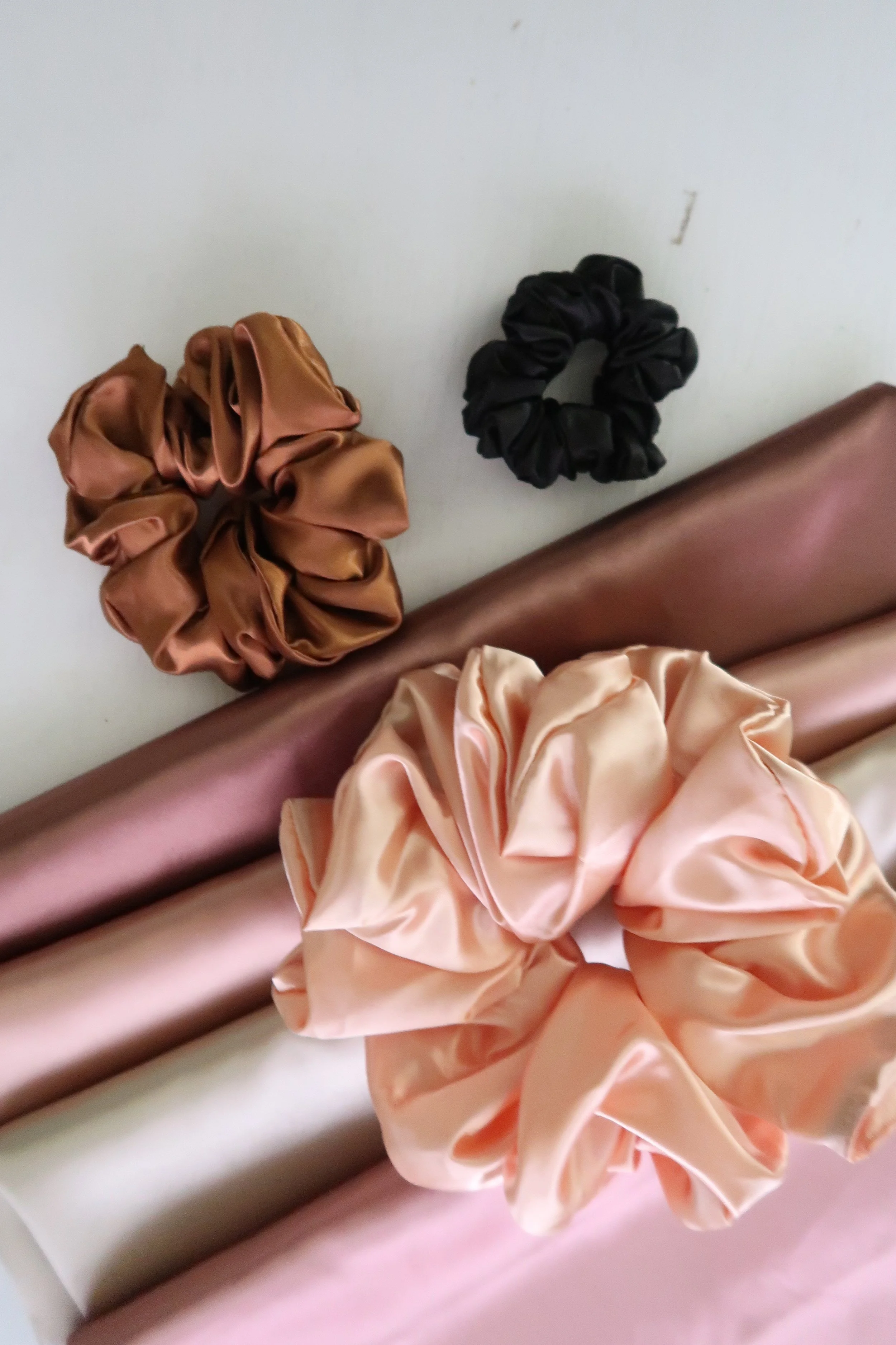 Close-up of two satin scrunchies on a white surface, one in light brown and the other in black, with a pink satin fabric and a metallic surface nearby.