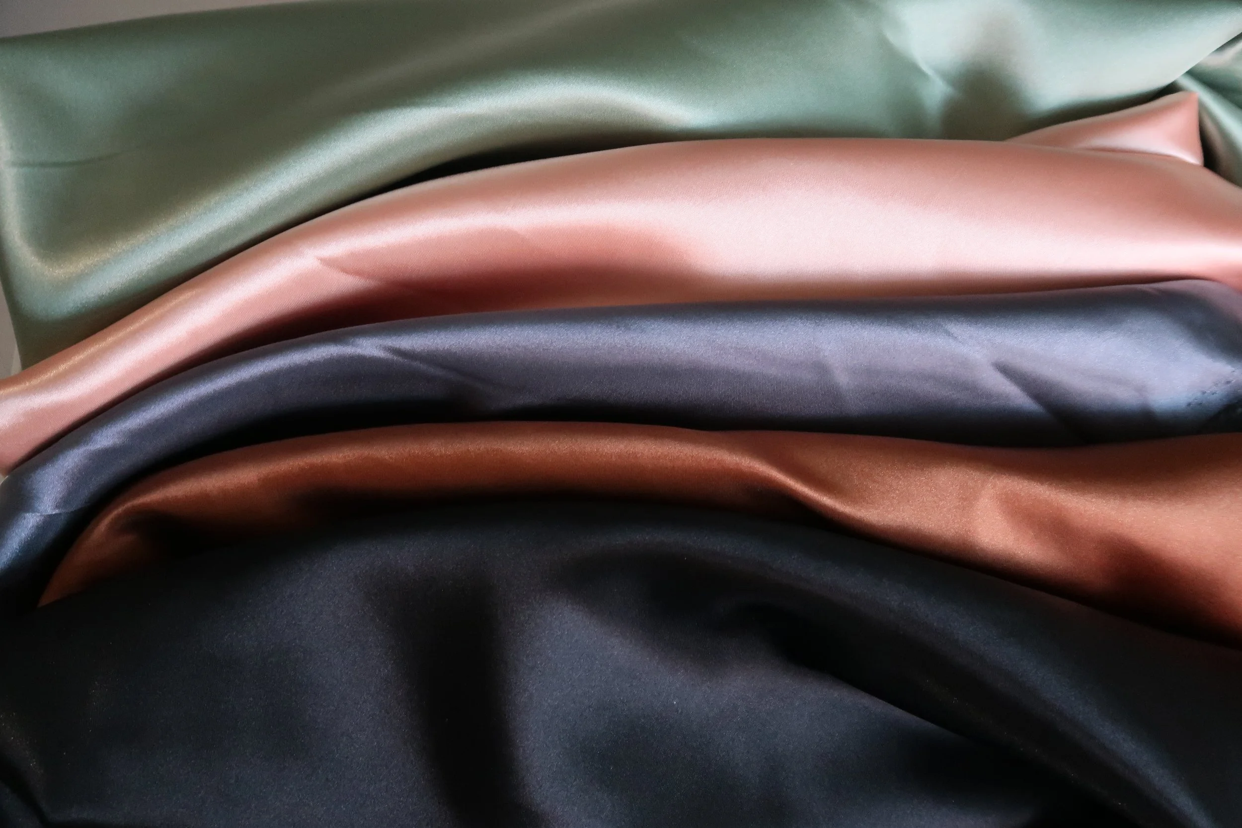 Stacked satin fabric in green, pink, gray, copper, and black colors.