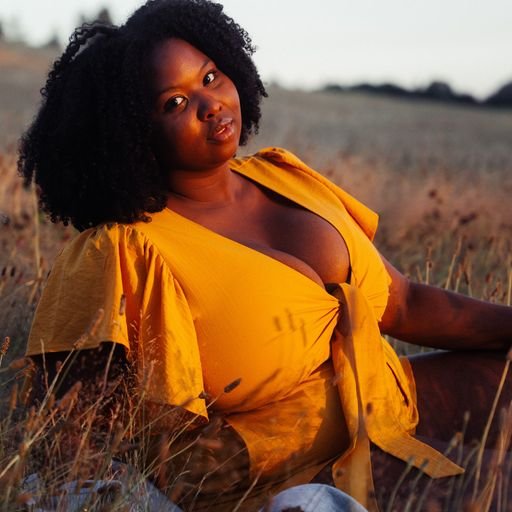 A woman with curly black hair wearing a yellow dress, sitting in a field of tall grass at sunset.
