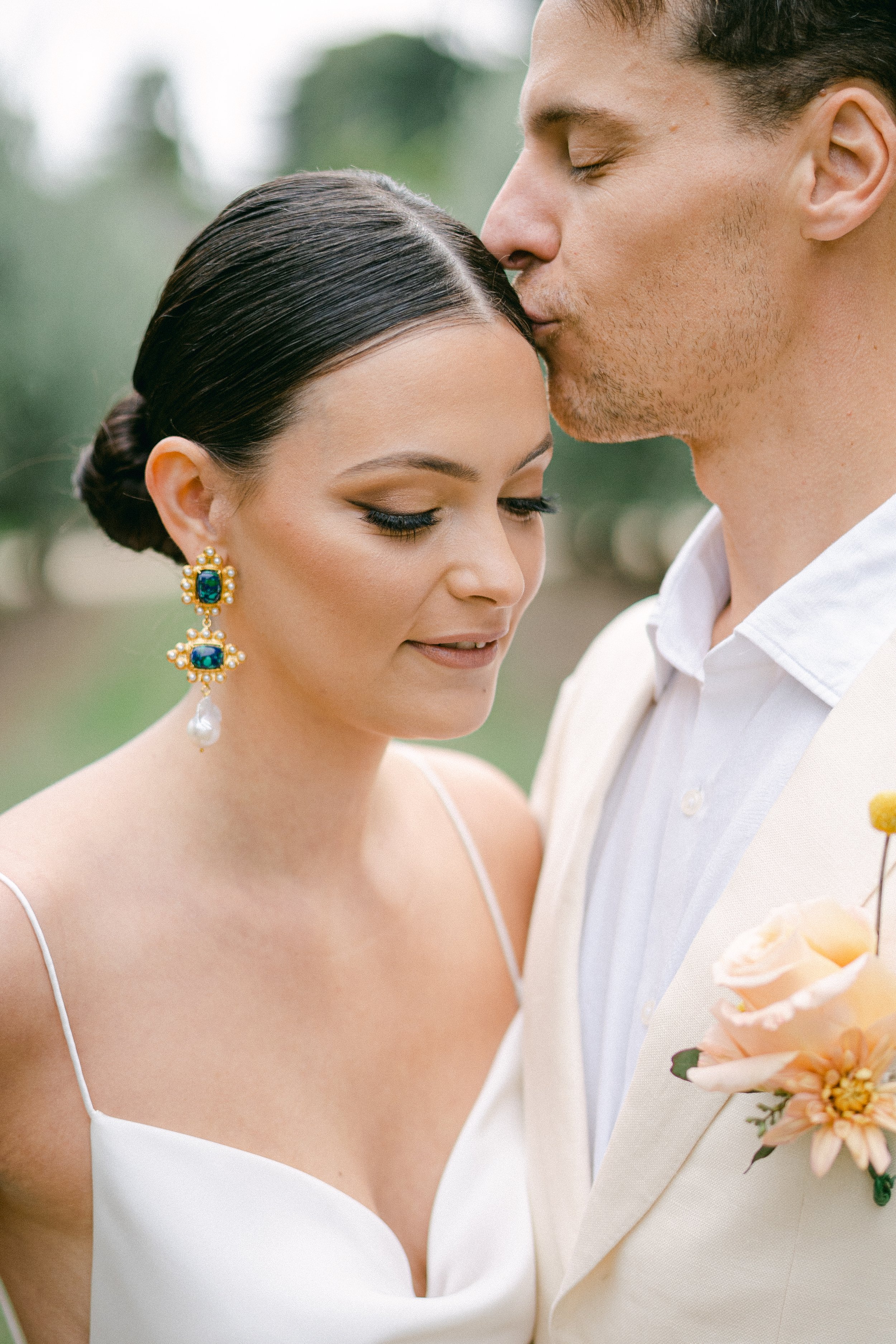 Bride and groom embracing, groom kissing bride's forehead, outdoors, bridal attire and bouquet.