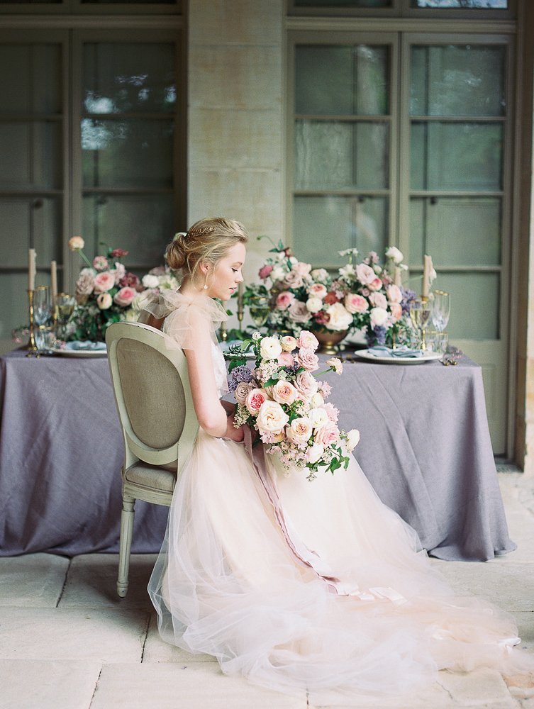 Bride sitting at a table with a large bouquet and floral arrangement in a soft, romantic setting.