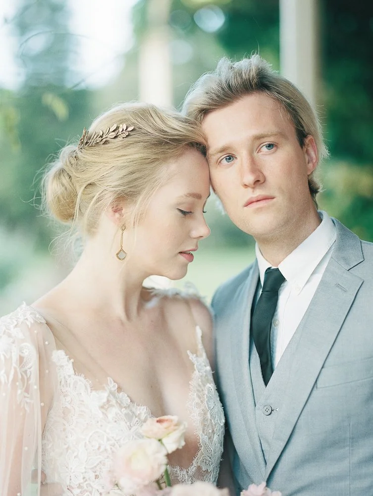 A romantic couple in wedding attire, with the woman wearing a lace dress and holding a bouquet, leaning against a man in a light gray suit with a vest and black tie. The setting is outdoors with a blurred leafy background.