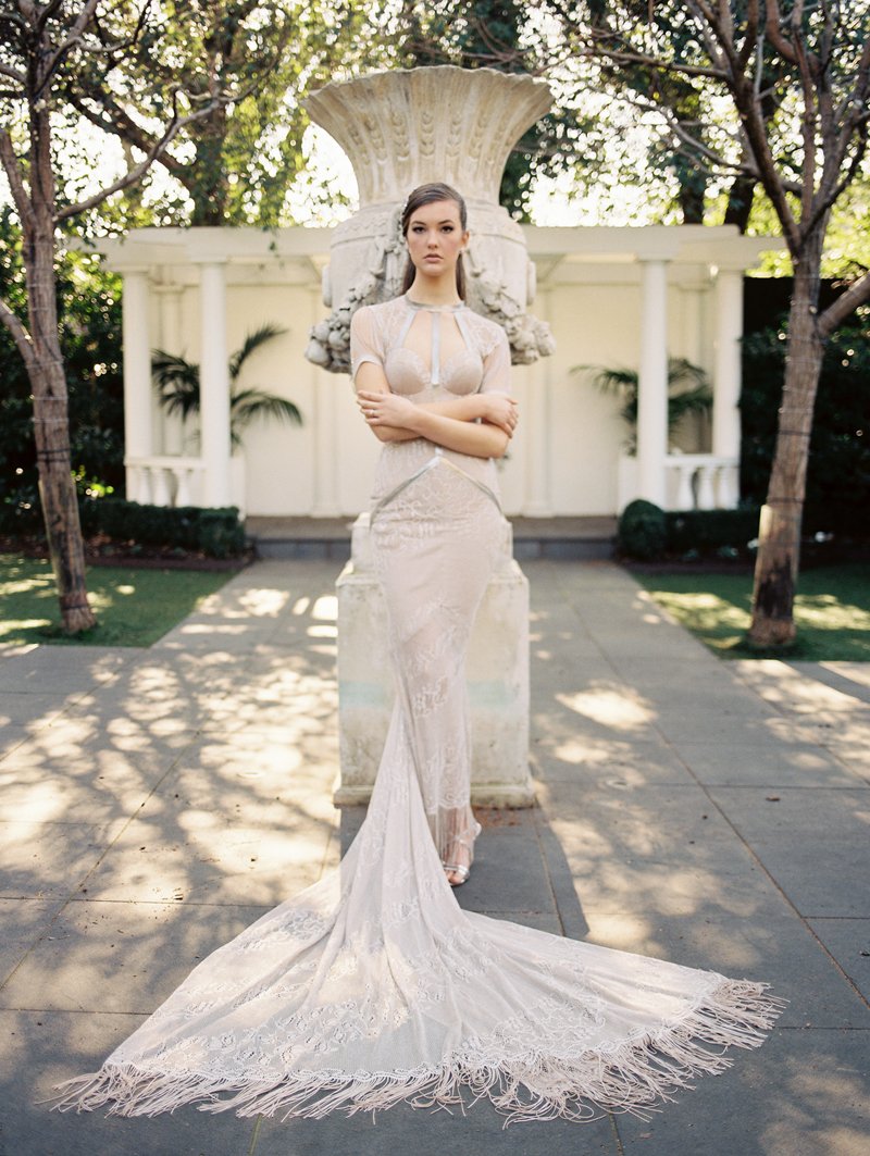 Woman in an elegant lace gown standing in front of a large stone urn in a garden setting with trees and white columns.