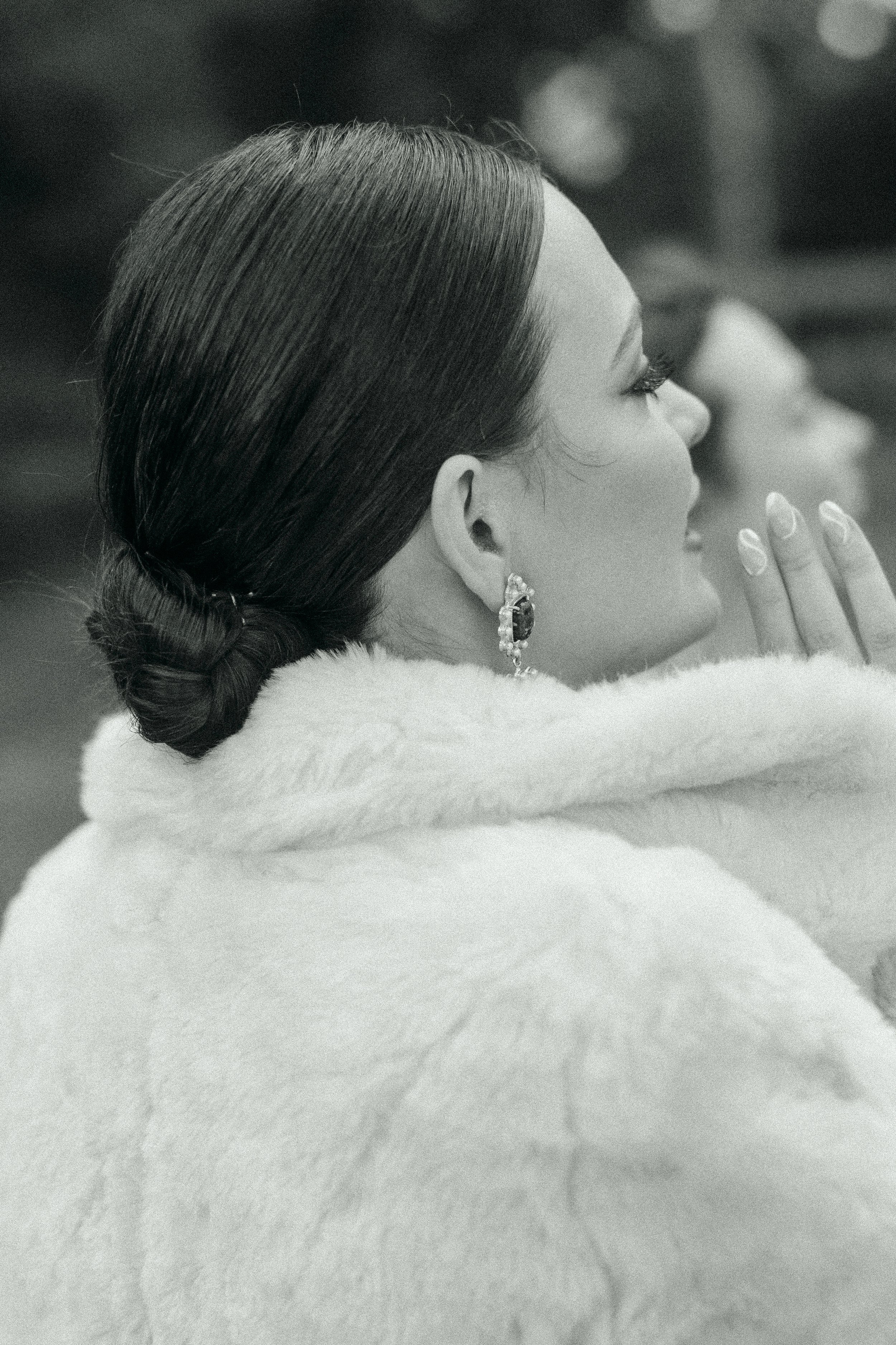 Back view of a person with a sleek hairstyle wearing a white fur coat, adorned with sparkly earrings, hands clasped together.