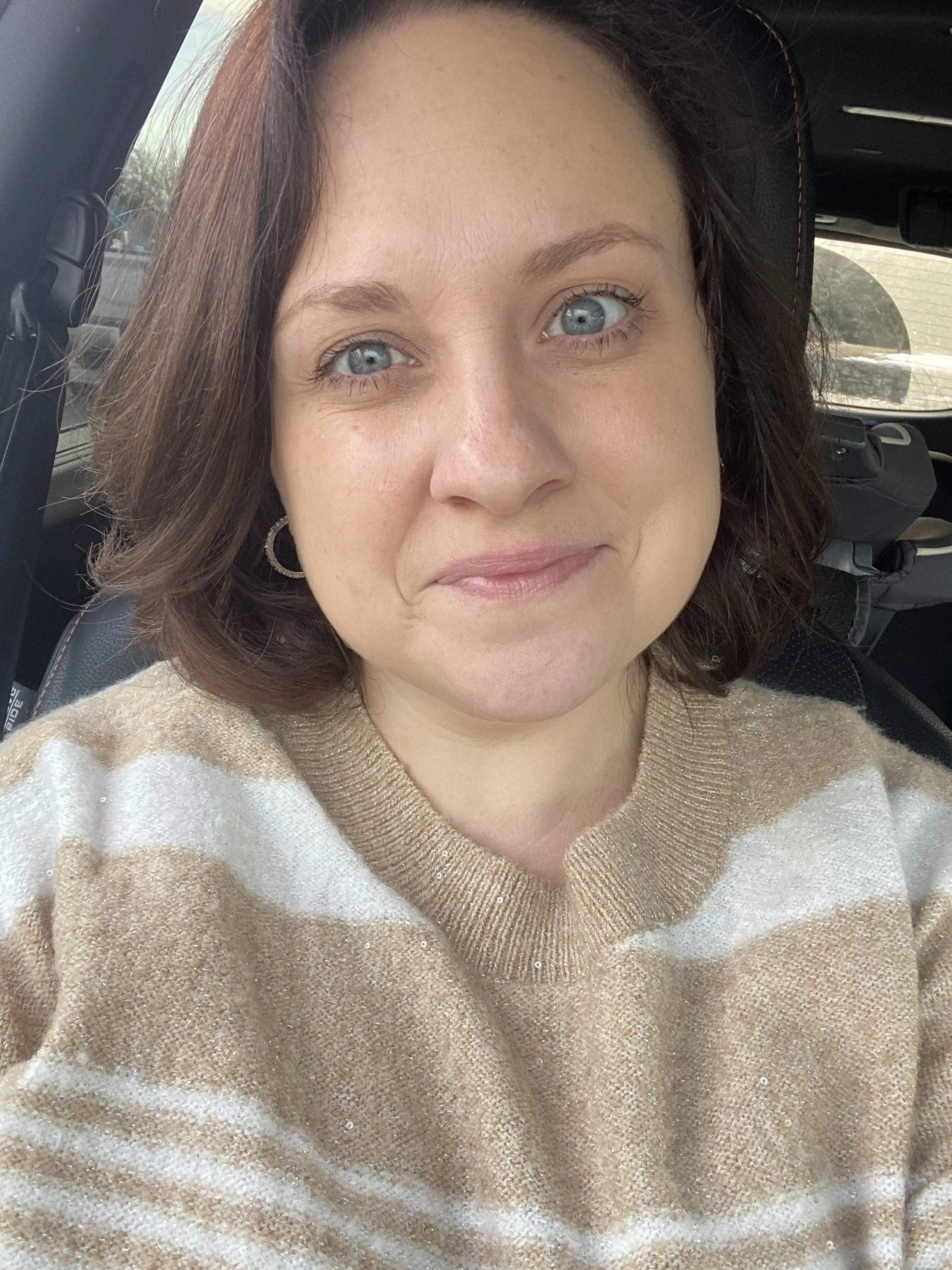 A woman with short brown hair and blue eyes wearing a tan and white striped sweater, sitting inside a vehicle.