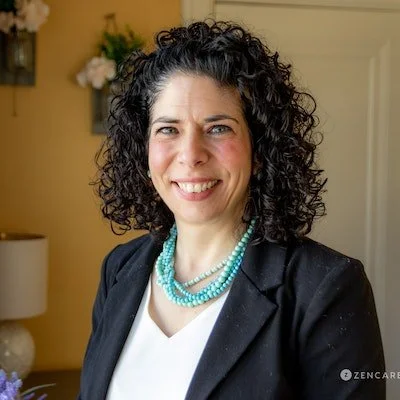 Woman with curly dark hair wearing a black blazer and turquoise necklace, smiling indoors.