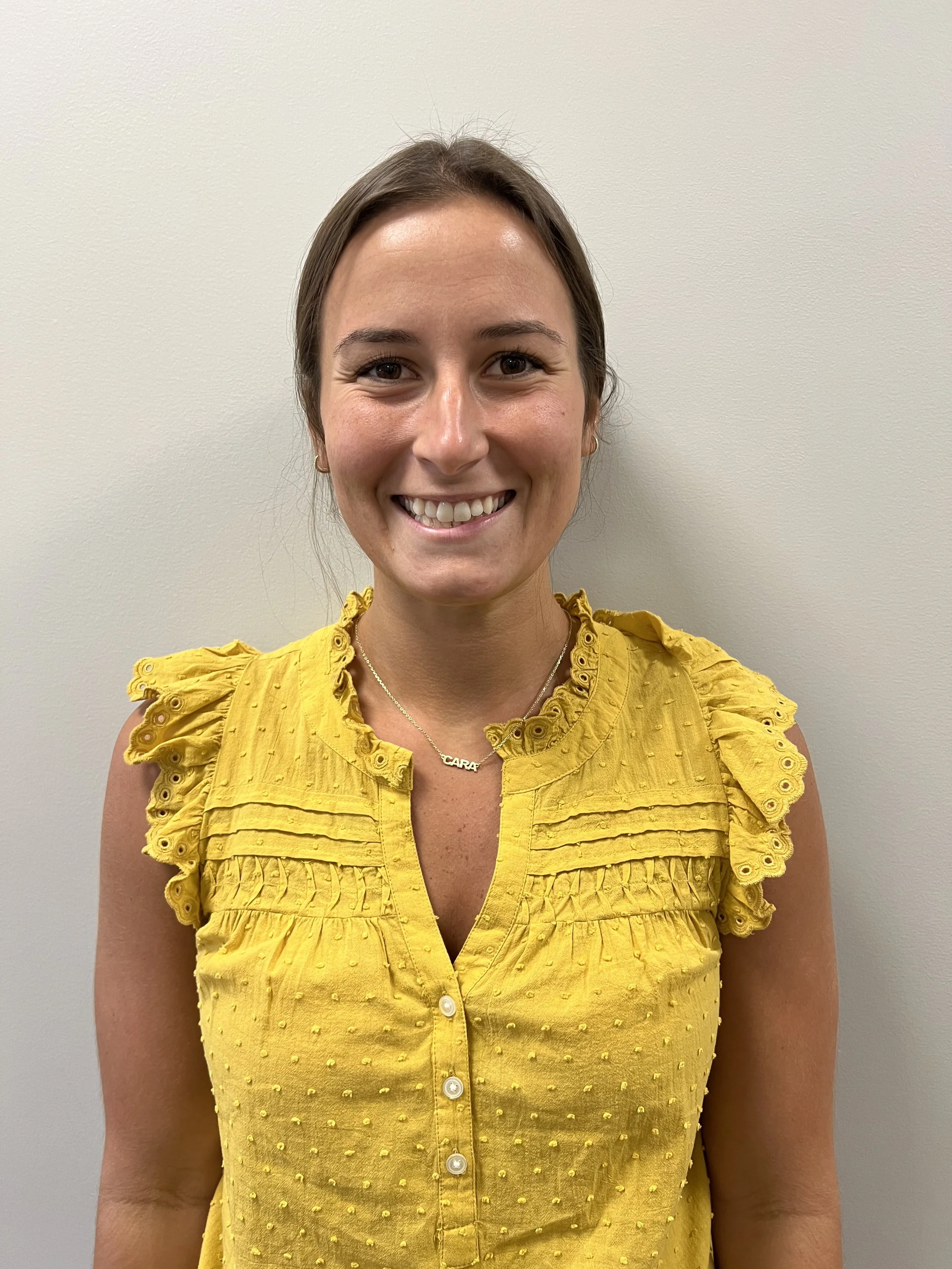 A woman with brown hair smiling, wearing a yellow sleeveless blouse with ruffled details and a name necklace that reads 'CARA' stands against a plain light-colored wall.