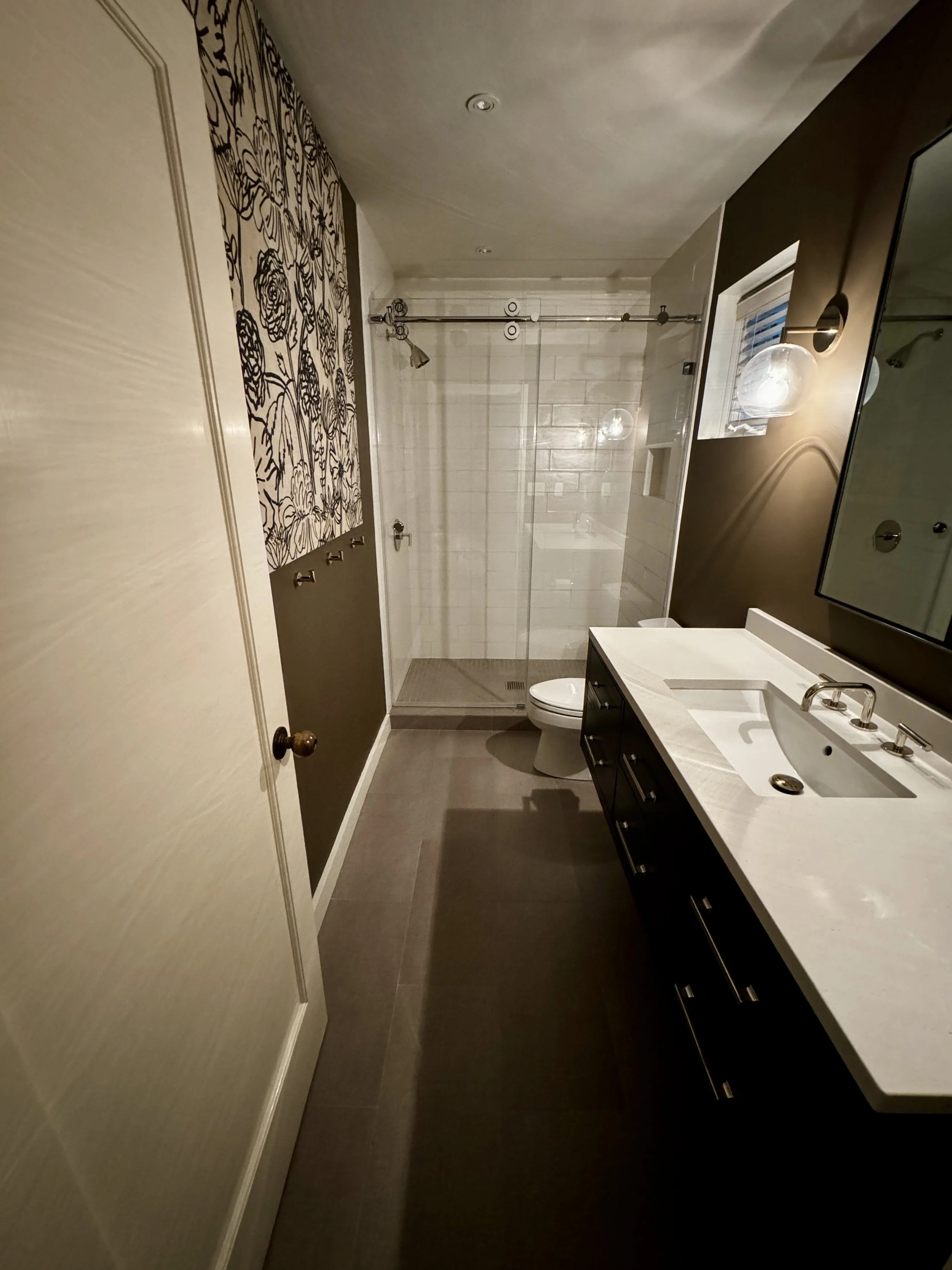 A modern bathroom featuring a dark vanity with a white countertop, a rectangular sink, a large mirror, a brushed nickel faucet, a toilet, and a walk-in shower with a glass door, white tiles, and a sliding shower curtain rod.