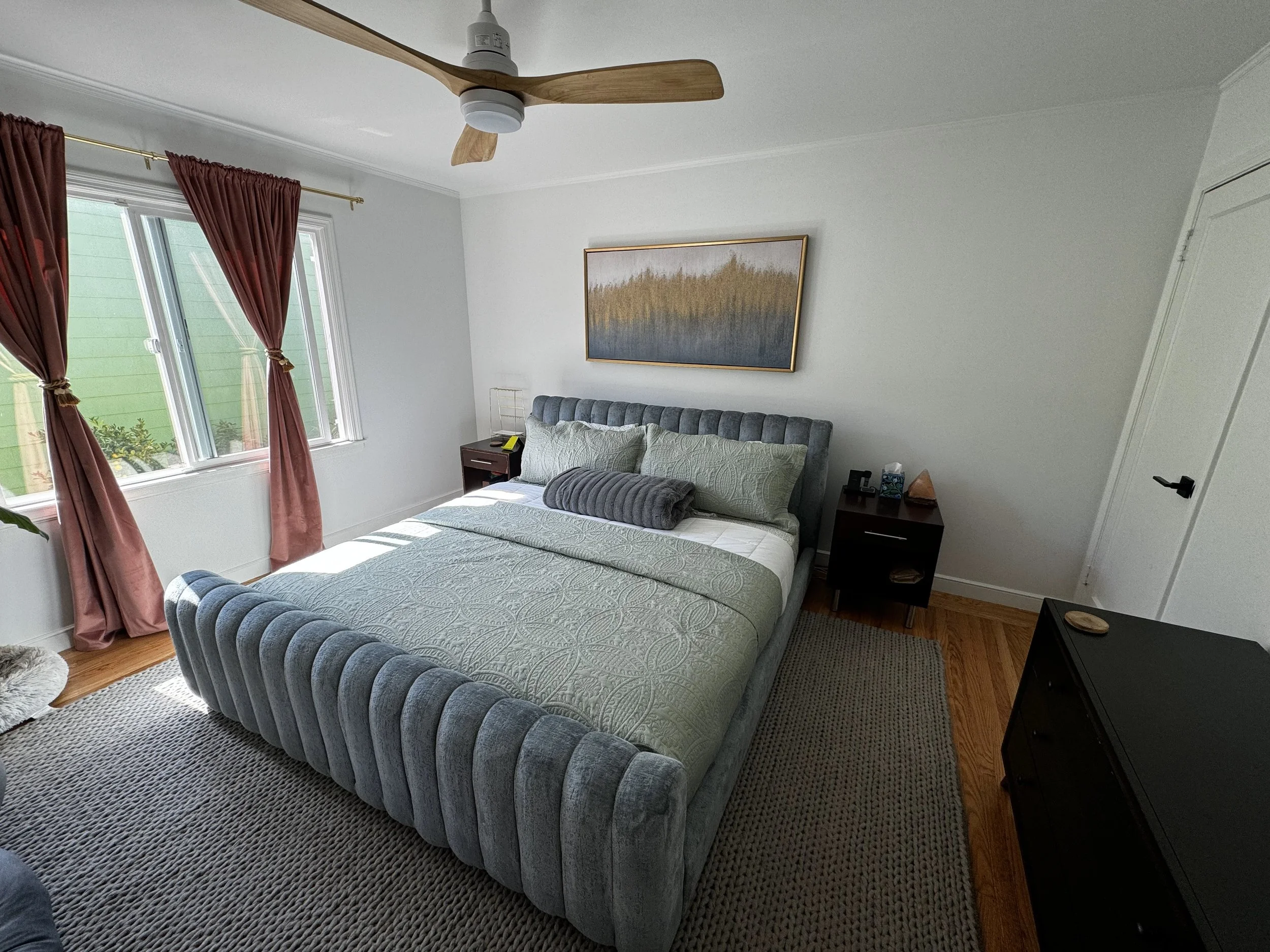 A bedroom with a bed, pink curtains, a window, a ceiling fan, a piece of artwork on the wall, and two nightstands, one with a lamp and the other with a tissue box and other items.