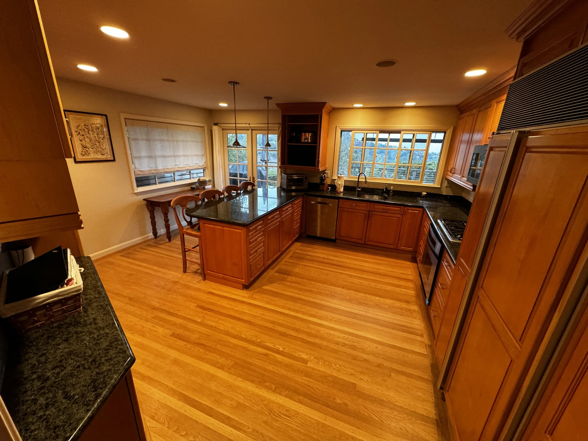 Interior view of a kitchen with wooden cabinetry, black countertops, a large window, and a dining area with sliding glass doors leading outside.