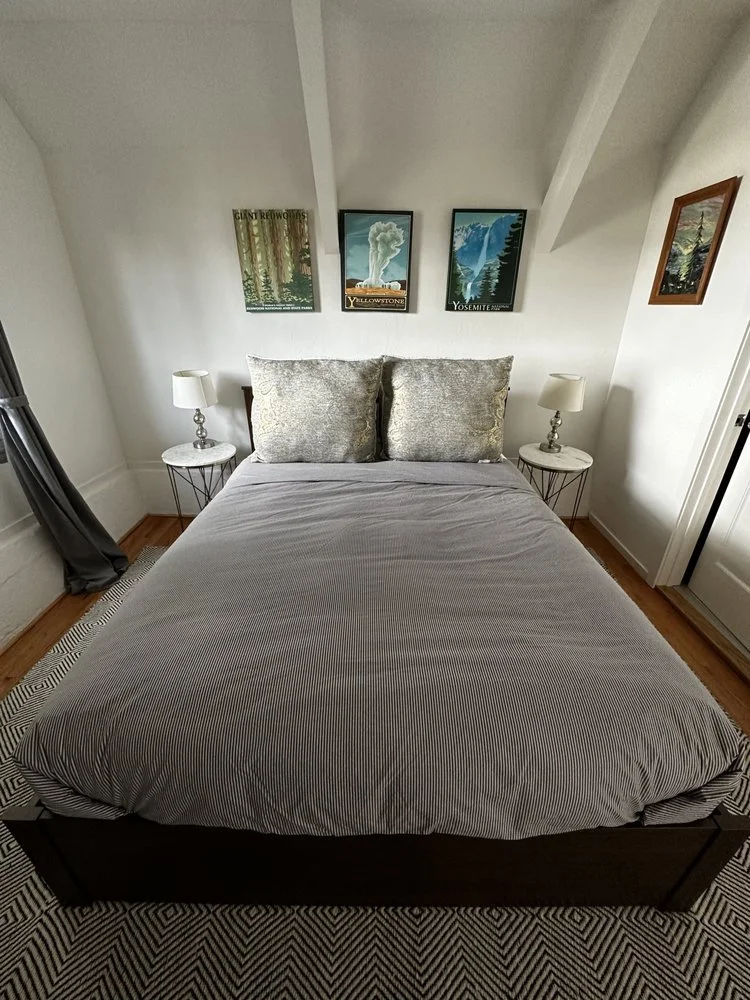 A bedroom with a bed, two pillows, two nightstands with lamps, and framed landscape artwork on the wall.