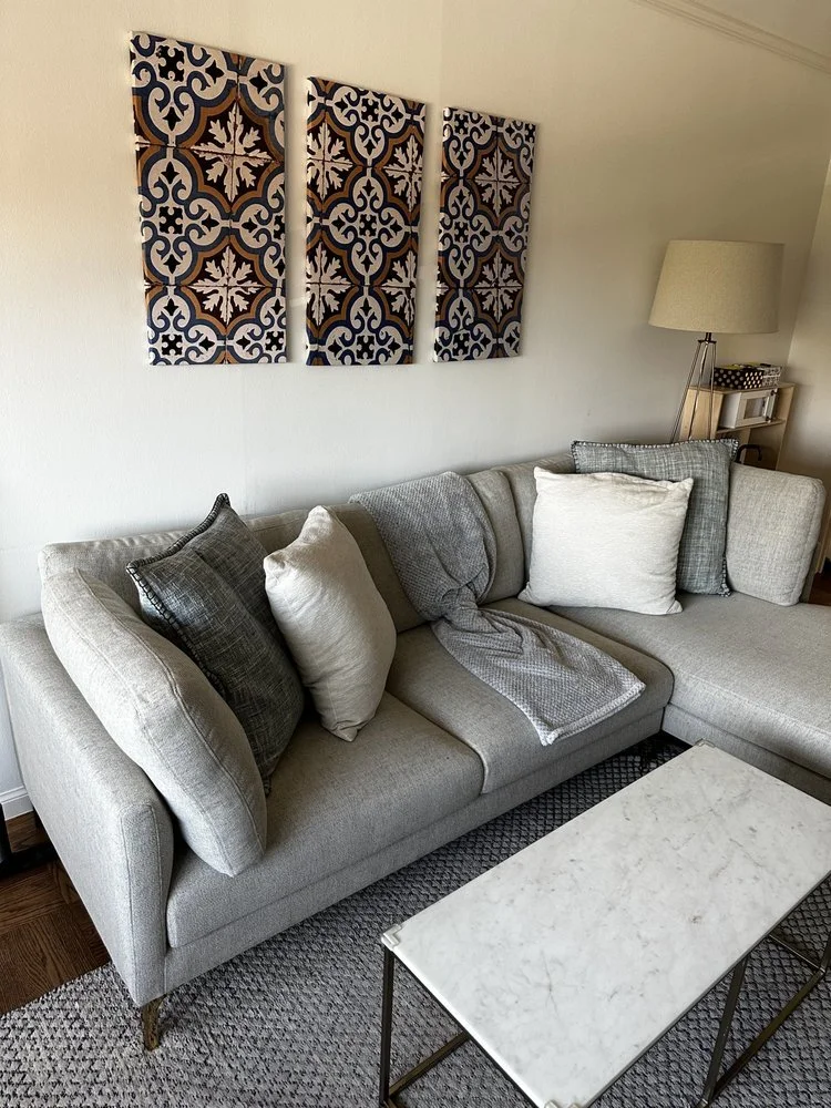 A living room with a gray sectional sofa decorated with various gray and white pillows, set against a white wall with three decorative patterned wall panels. A small white marble coffee table with a metal frame sits in front of the sofa, and a floor lamp with a shade is visible in the corner.