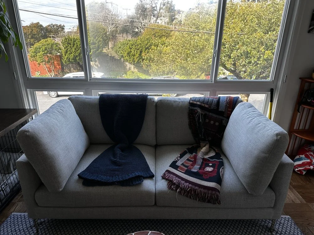 Light gray sofa in front of large window with trees outside, draped with two colorful blankets.
