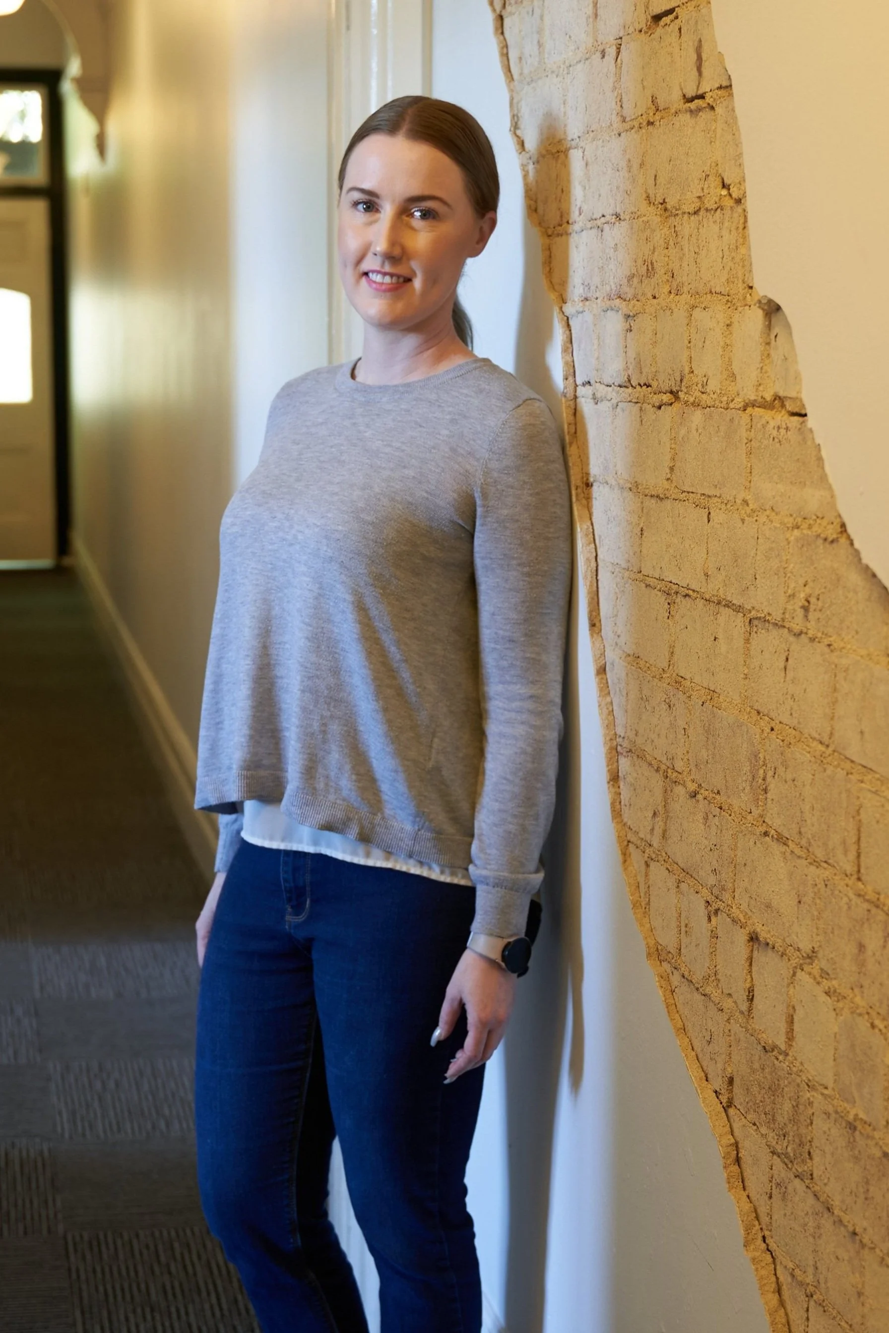 KK Stanley, A woman with brown hair tied back, wearing a gray sweater, dark blue jeans, and a wristwatch, standing and smiling against a wall with exposed bricks.
