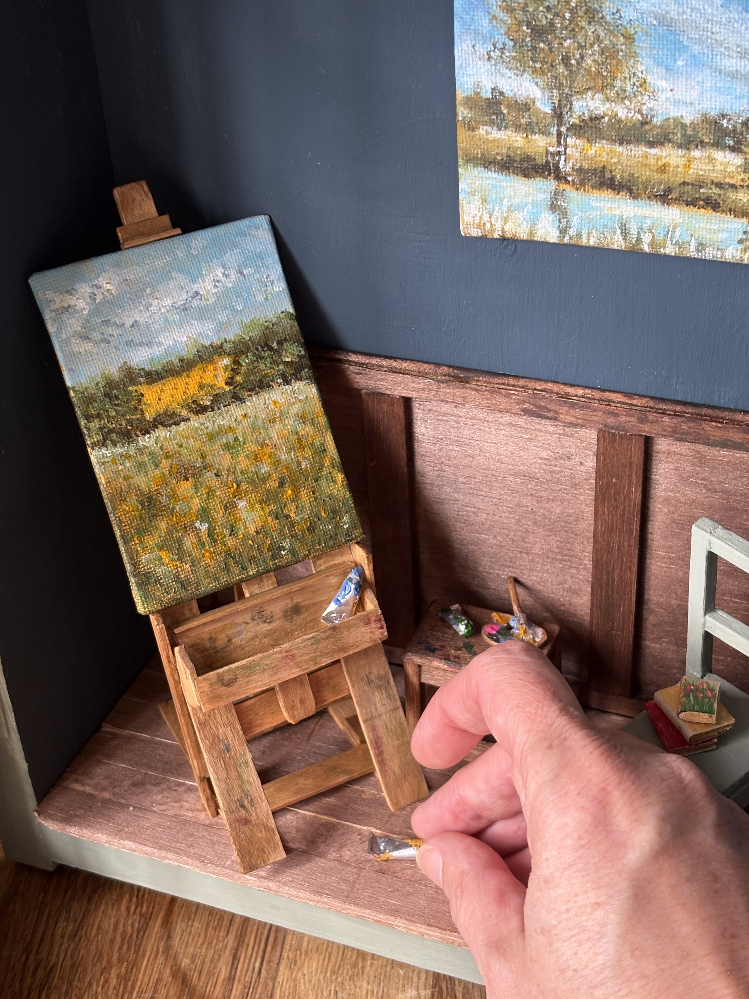 Hand touching miniature landscape artist studio diorama. Handcrafted wooden easel, table and chair. Landscape acrylic painting of corn field.