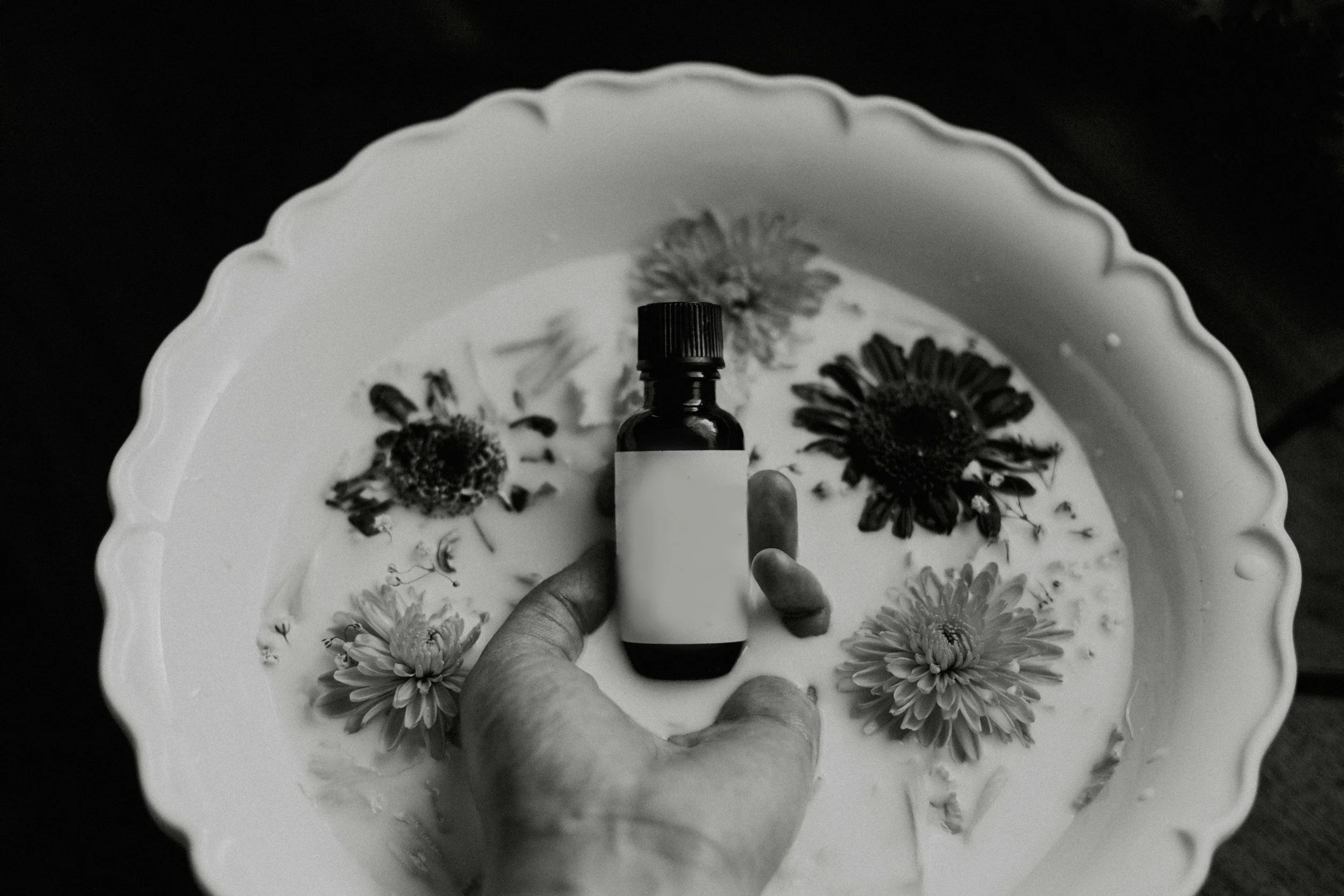 A hand holding a small bottle with a blank label and a cap, over a bowl filled with water, flowers, and a small stone, captured in black and white.