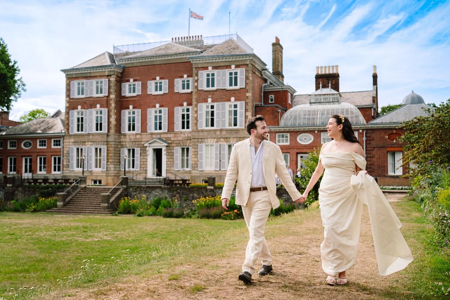 A bride and groom walk hand in hand with York House registry office in Twickenham 
visible in the background.