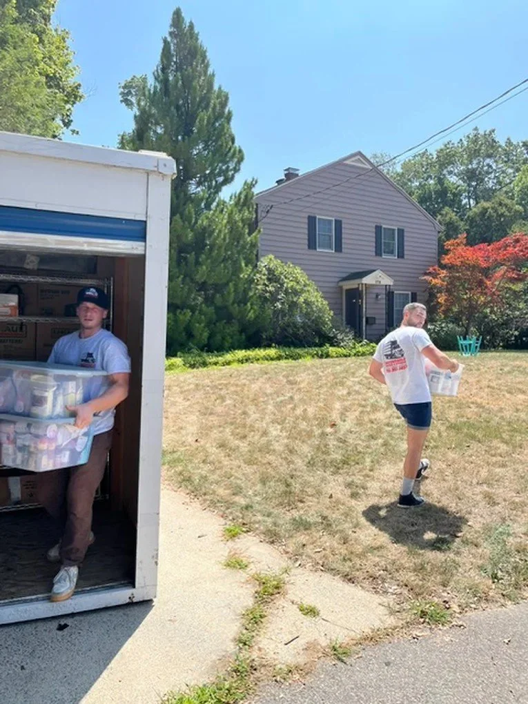 Two young men unloading items from a truck in a residential neighborhood on a sunny day. One is inside the truck carrying a clear plastic container, the other is outside holding a white box.