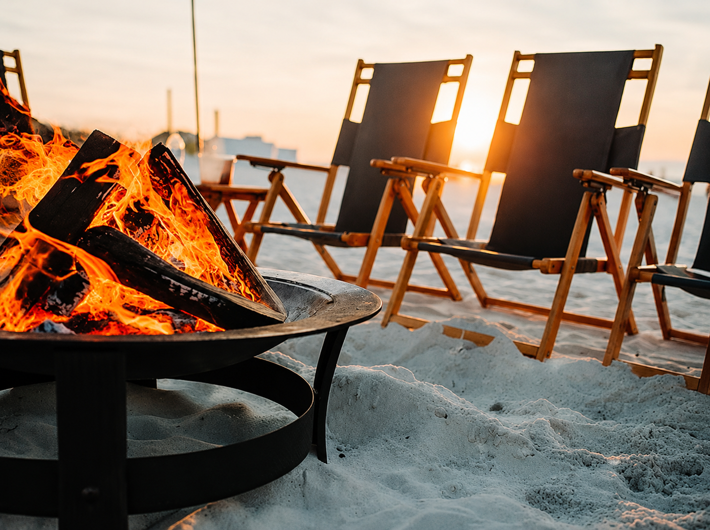 Beach scene with a firepit and three wooden deck chairs, sunset in the background.