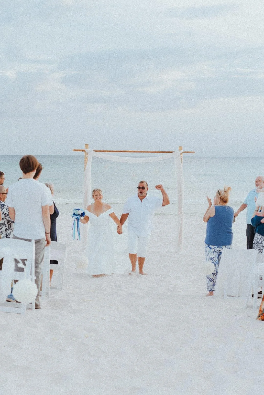 A beach wedding ceremony with a couple holding hands under an arch, surrounded by seated and standing guests, with the ocean and sky in the background.