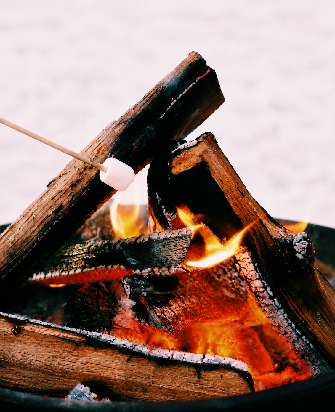 Close-up of a campfire with burning logs and flames, with a marshmallow on a stick roasting over the fire.