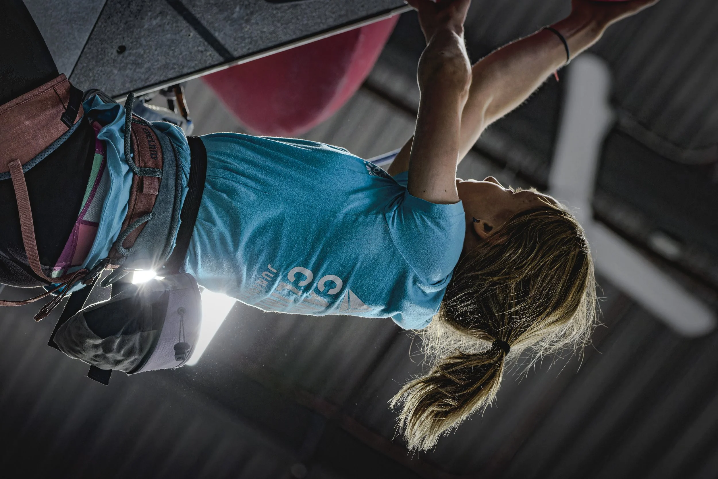 An indoor rock climber with a ponytail wearing a blue shirt and climbing harness, reaching for holds on a climbing wall.