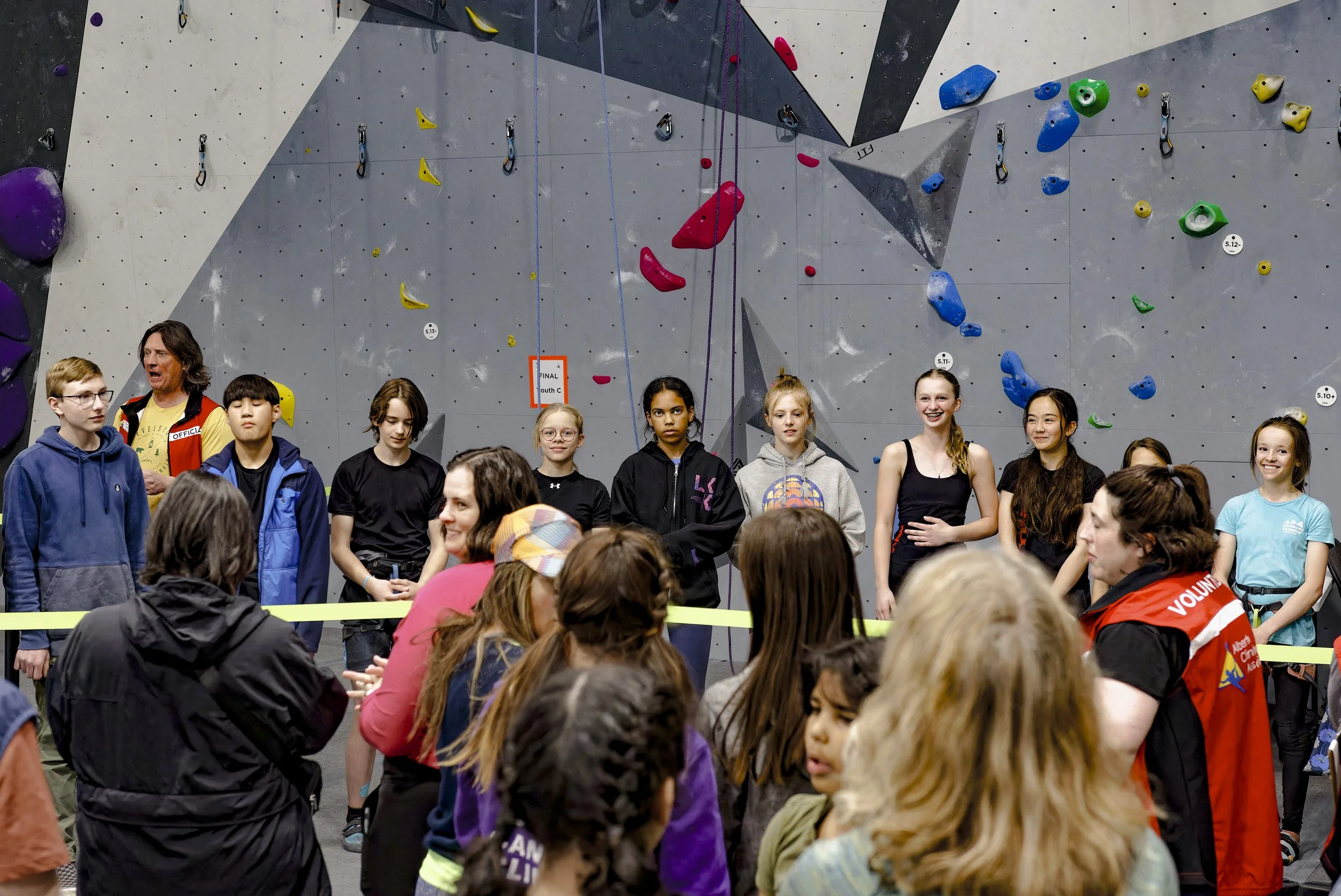 Group of young girls and adults standing in front of a climbing wall at an indoor rock climbing gym.