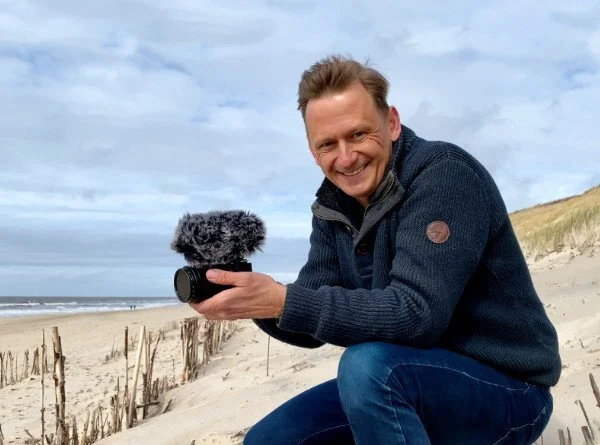 Mann in blauem Pullover hält Kamera mit Mikrofon an einem Strand.