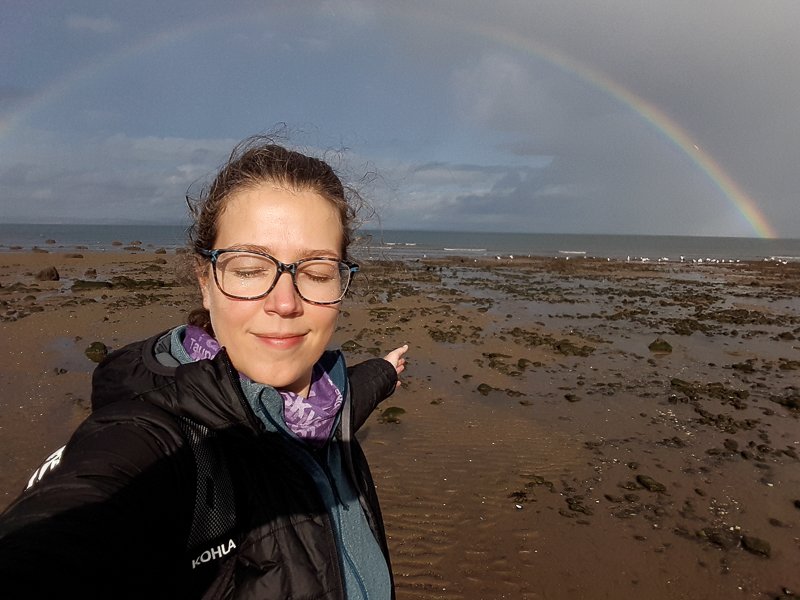 Lucie Aidart en randonnée méditation, sur la plage à Édimbourg en Écosse, un arc-en-ciel au-dessus d'elle.