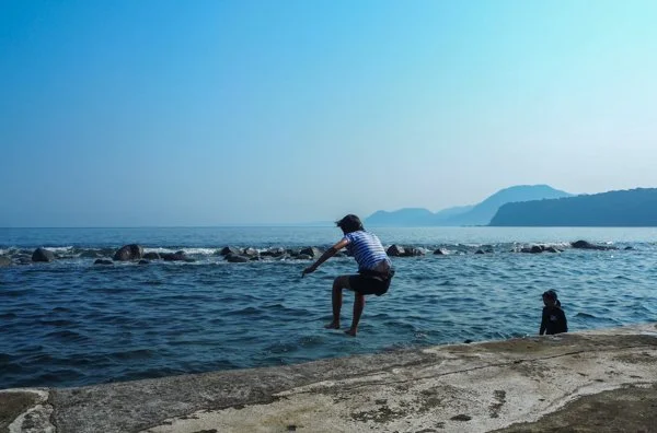 Child jumping into water at the beach with another child sitting nearby, ocean and mountains in the background under a clear sky.