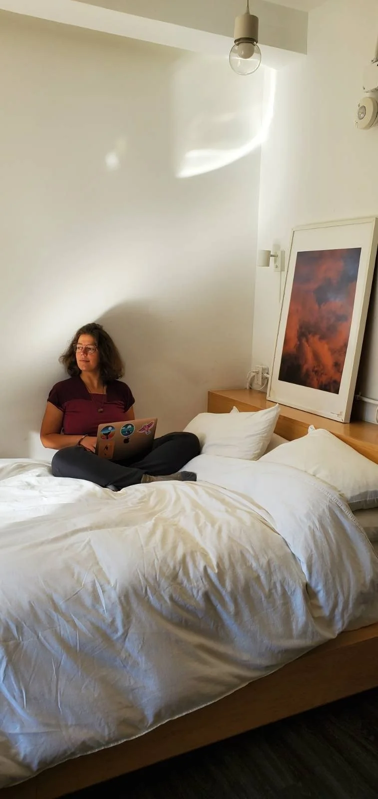 A woman sitting cross-legged on a bed using a laptop, with a minimalist room background featuring a large framed picture of clouds, a bedside table, and a ceiling light.