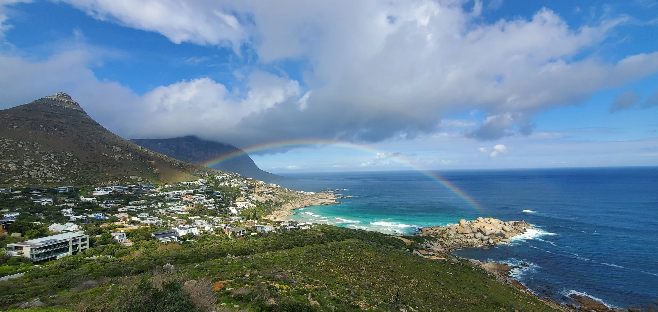 A coastal landscape with a rainbow arching over the ocean, hillside residential buildings, a mountain, and a partly cloudy sky.