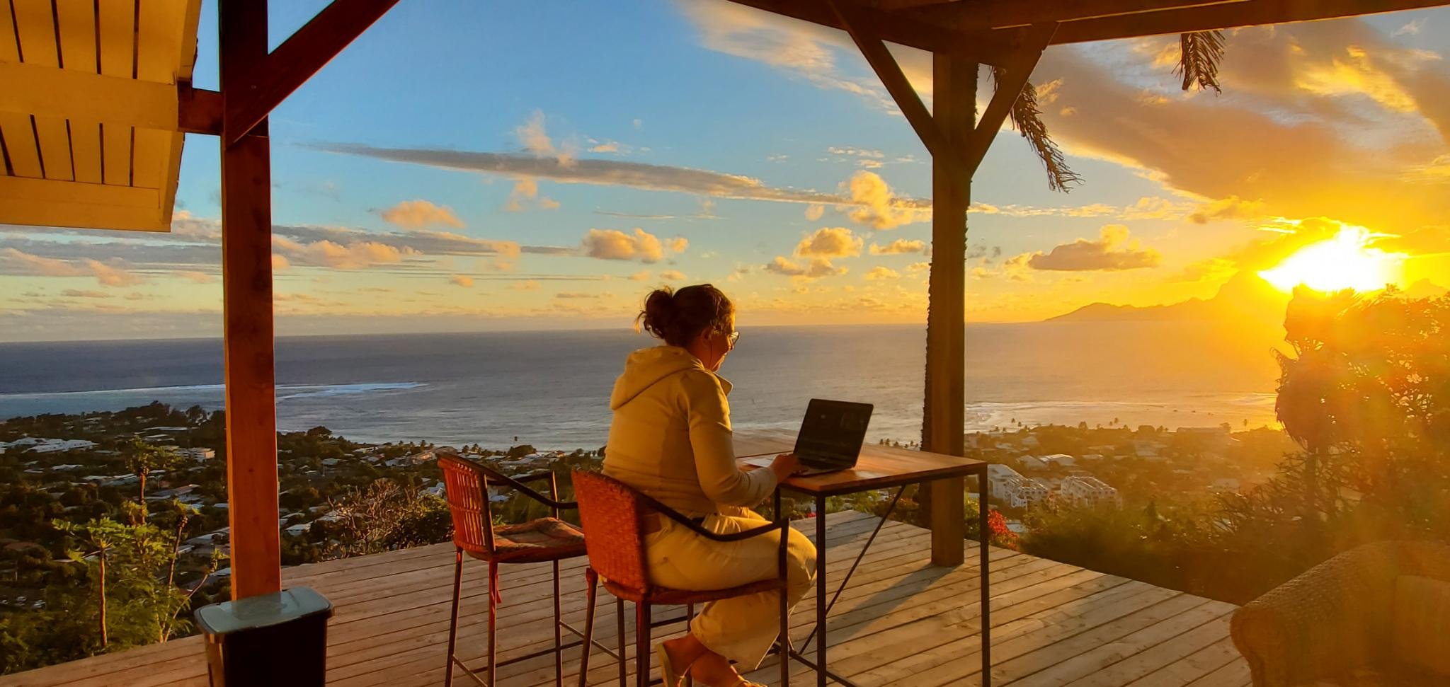A writer sat at a computer, on a wooden terrace, in Tahiti, with a view of the Pacific Ocean and sunset on Moorea island