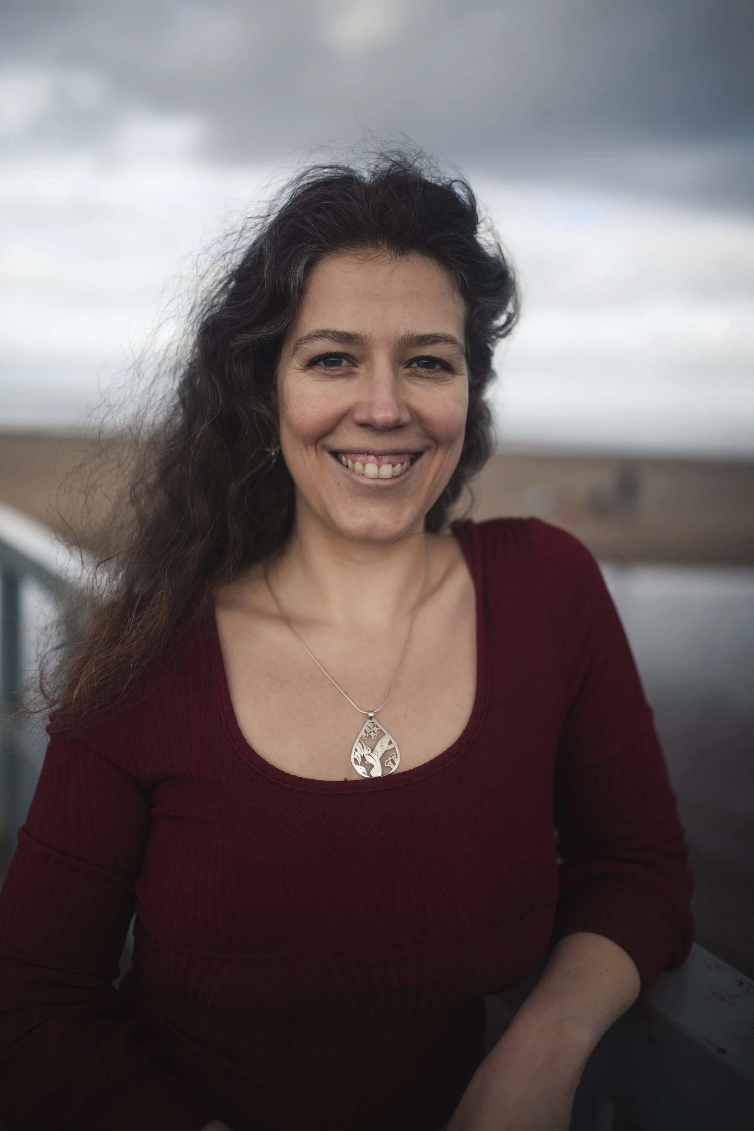 A woman wearing a maroon long-sleeve shirt and a silver necklace, smiling outdoors with a cloudy sky and landscape in the background.