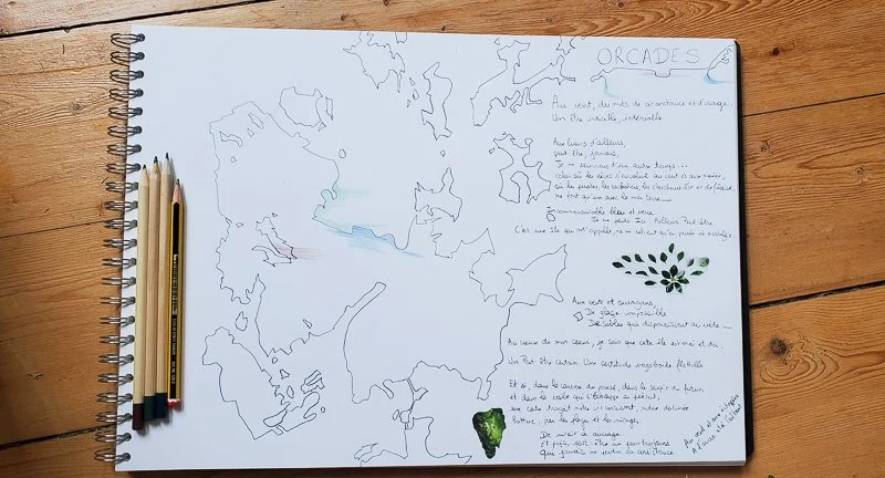 Open sketchbook with a handwritten map outline and notes, placed on a wooden surface, with four colored pencils on the left side.